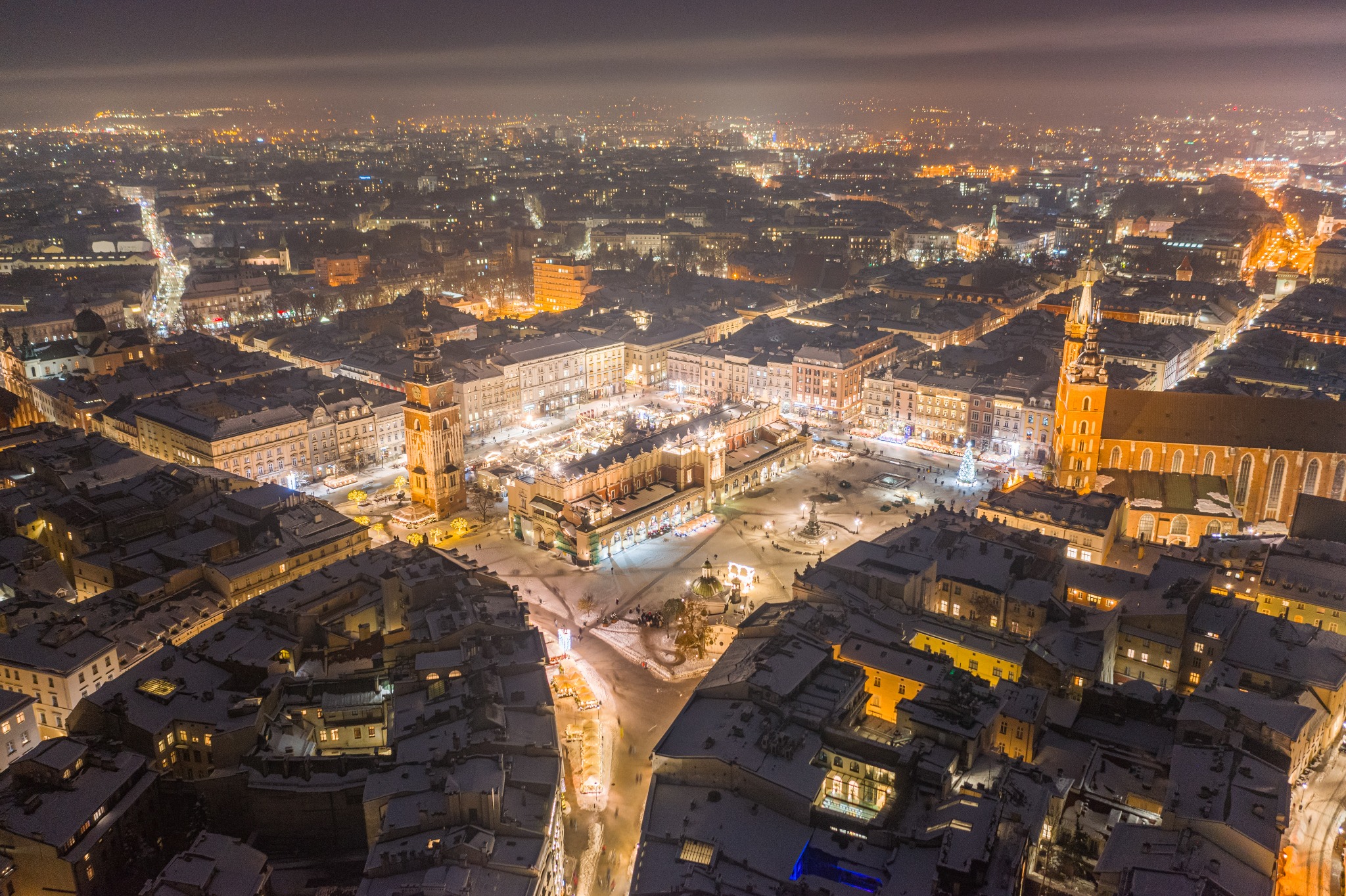 Nocny widok z lotu ptaka na ośnieżony Rynek Główny w Krakowie, oświetlony budynki Sukiennic, Kościół Mariacki i okoliczne kamienice, z widoczną choinką na placu i światłami miasta w tle.