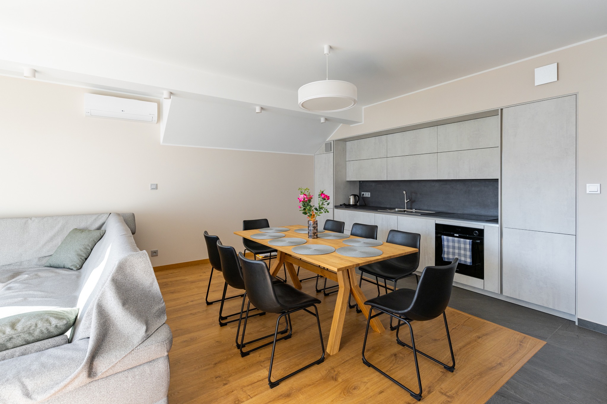 Jasne wnętrze mieszkania w Bielsko-Białej z drewnianym stołem, czarnymi krzesłami, szarą sofą i minimalistyczną kuchnią z szarymi szafkami oraz kwiatami w wazonie na stole.
