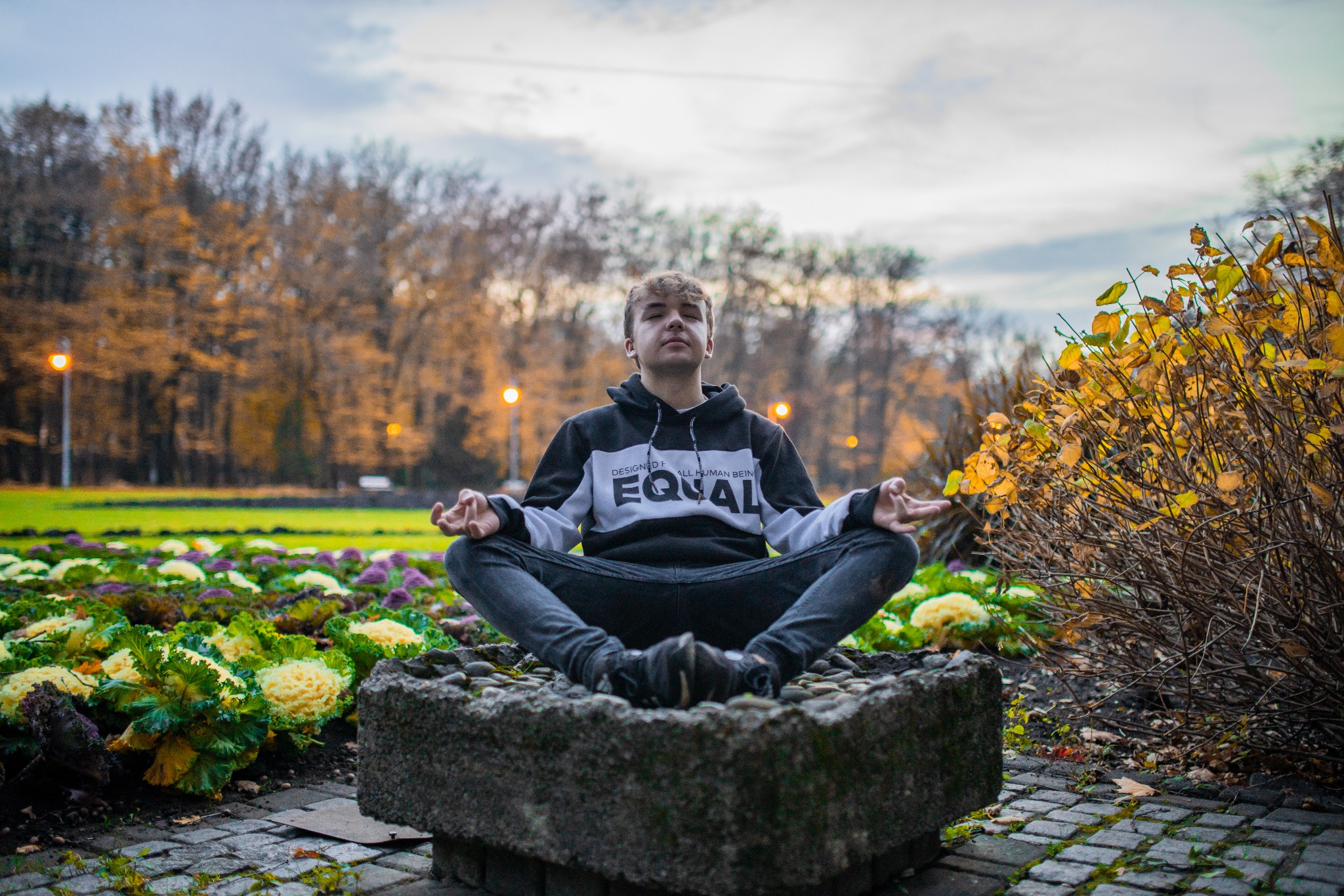 Młody mężczyzna w bluzie z kapturem siedzi w pozycji lotosu na kamiennym postumencie w parku, z rabatami kwiatowymi i jesiennymi drzewami w tle.
