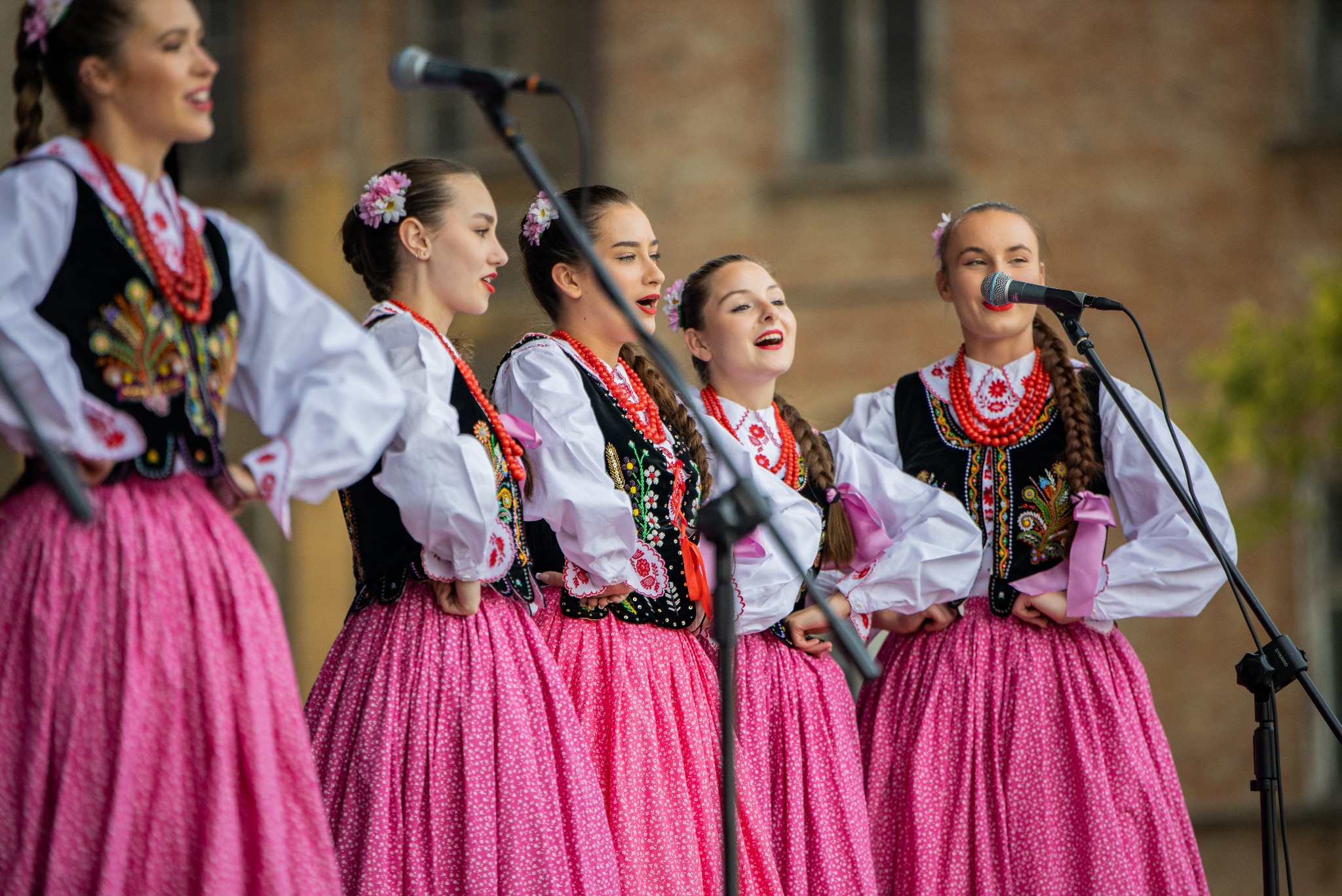 Występ zespołu ludowego w Bielsku-Białej: pięć młodych kobiet w tradycyjnych strojach śpiewa do mikrofonów, ubrane w białe bluzki, czarne haftowane kamizelki i różowe spódnice.