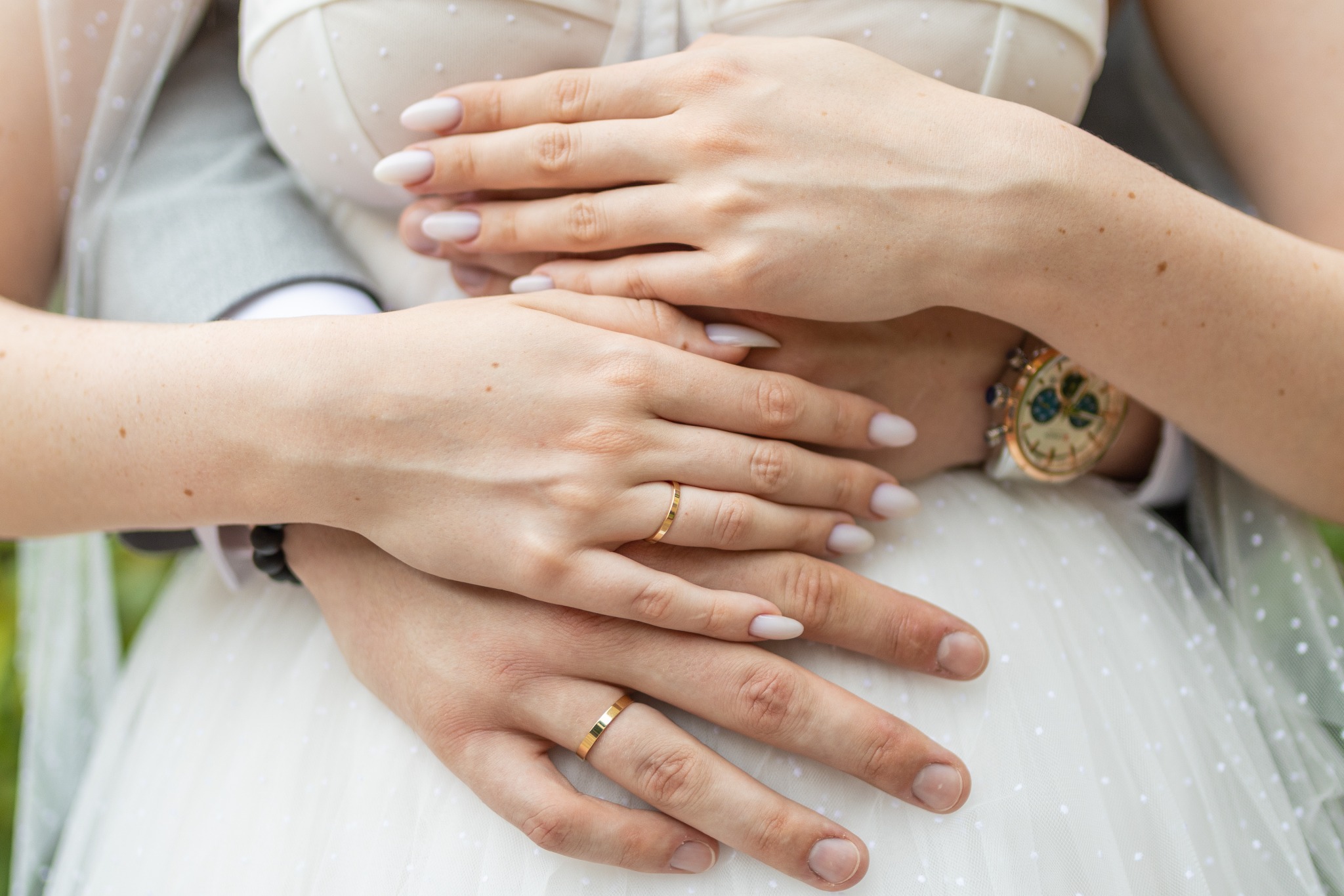 Dłonie pary młodej z obrączkami na tle sukni ślubnej i marynarki, widoczny zegarek męski z tarczą w odcieniach beżu i złota.