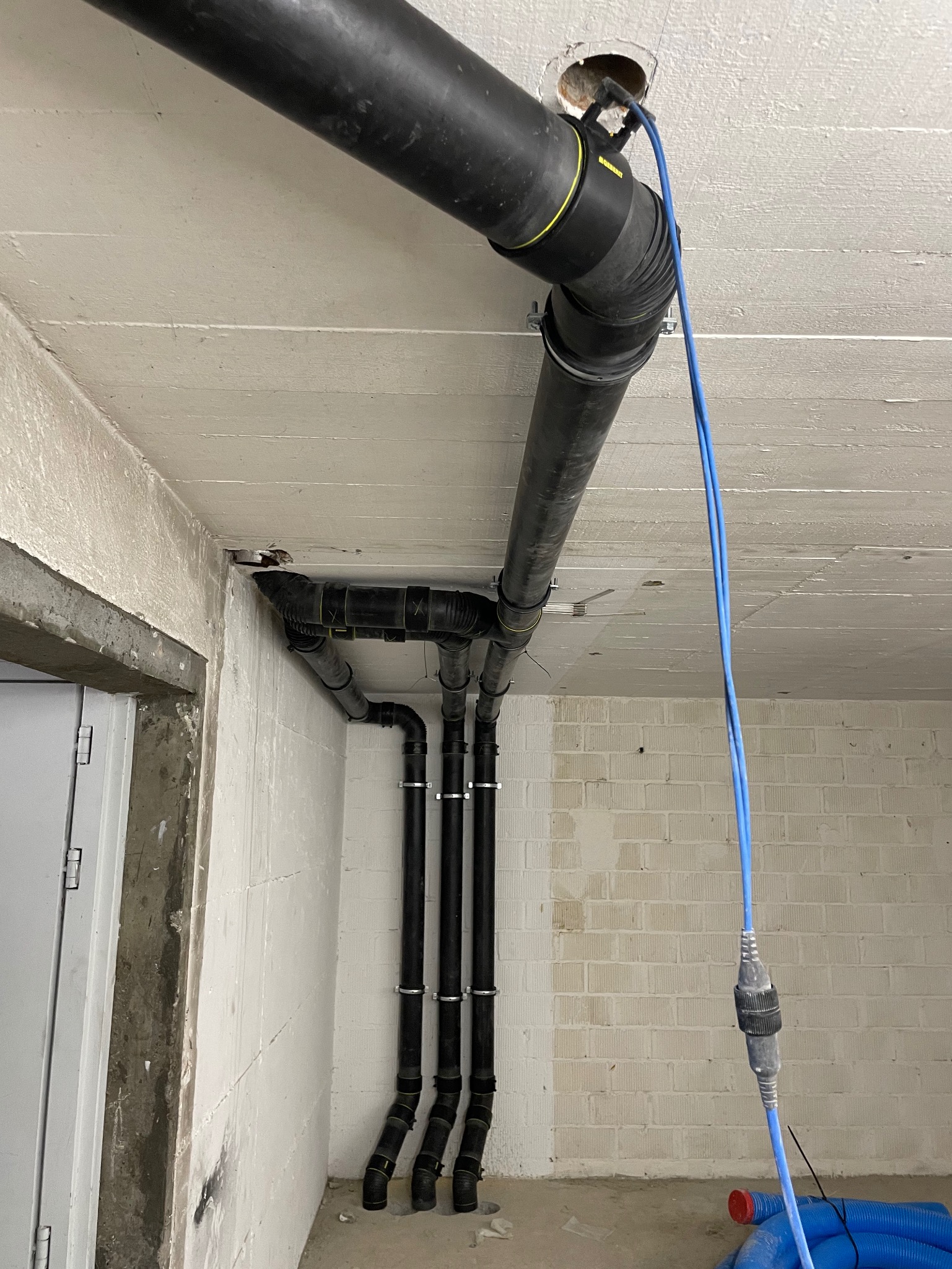 Czarna rura kanalizacyjna z żółtym pierścieniem przechodząca przez otwór w betonowym suficie, widoczne trzy pionowe rury z obejmami na tle ściany z białego pustaka, niebieski przewód zwisający...