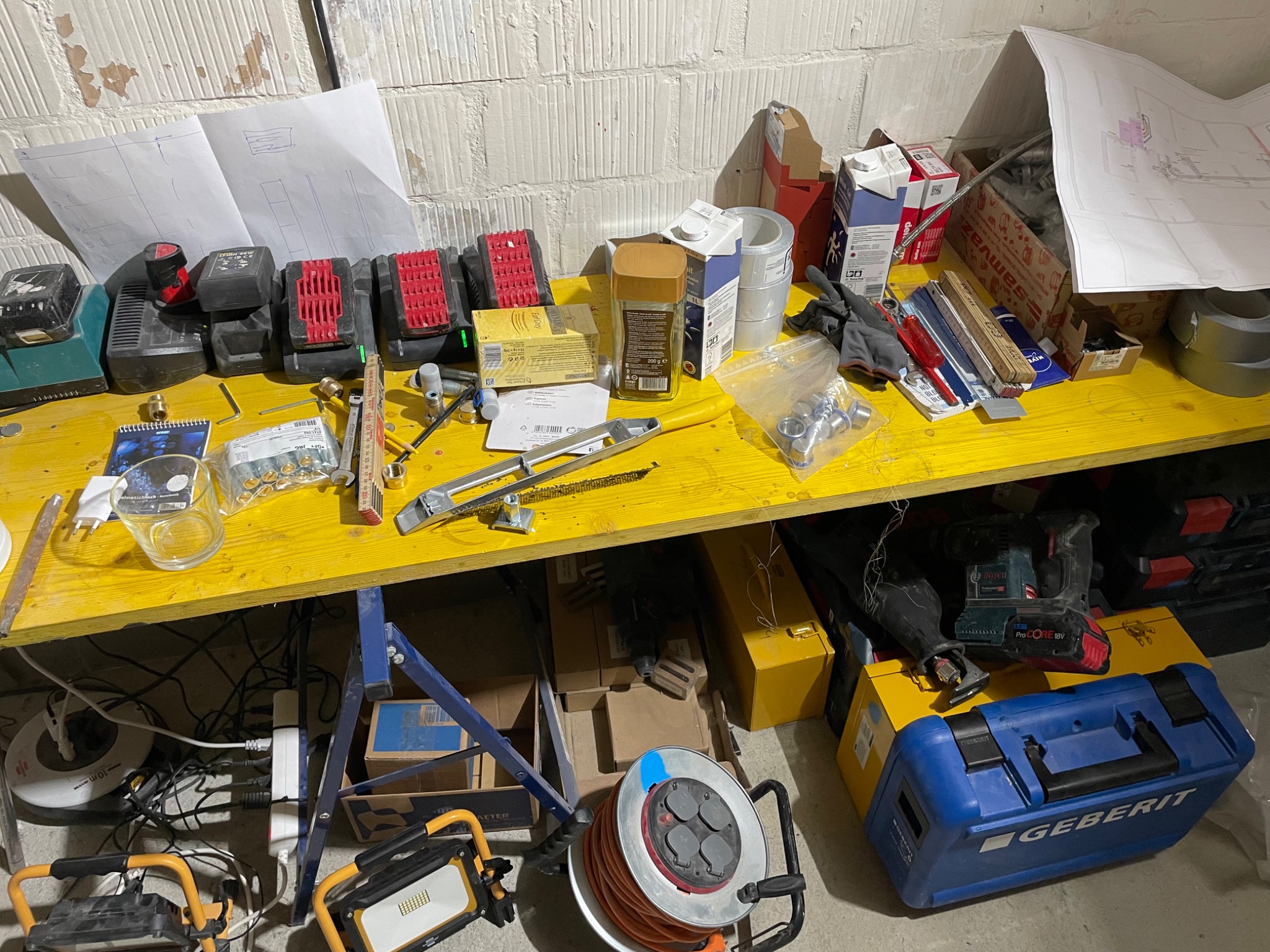 Żółty stół roboczy z narzędziami hydraulicznymi, bateriami do elektronarzędzi, kawą, przedłużaczem, skrzynką narzędziową Geberit i planami instalacji na tle ściany z cegieł.