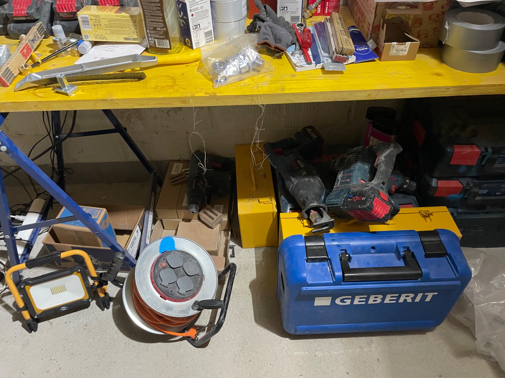 Warsztatowy bałagan: żółty stół roboczy z narzędziami, rurami, taśmami i pudełkami, pod nim skrzynka narzędziowa Geberit, zwijany przedłużacz i lampa warsztatowa.