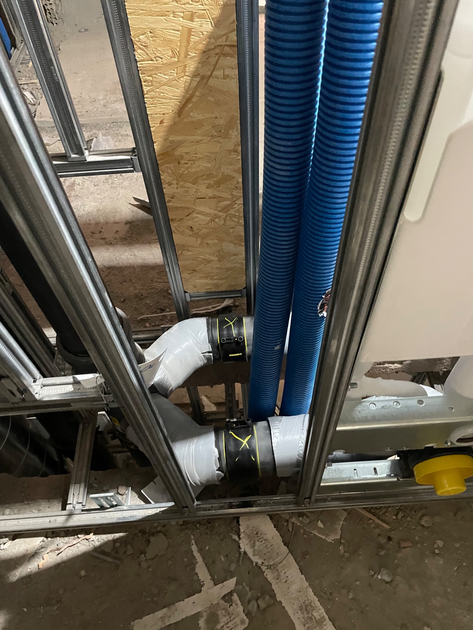 Szare rury kanalizacyjne z żółtymi oznaczeniami, niebieskie rury wentylacyjne i stelaż podtynkowy w trakcie montażu, widoczne elementy konstrukcji ścian.