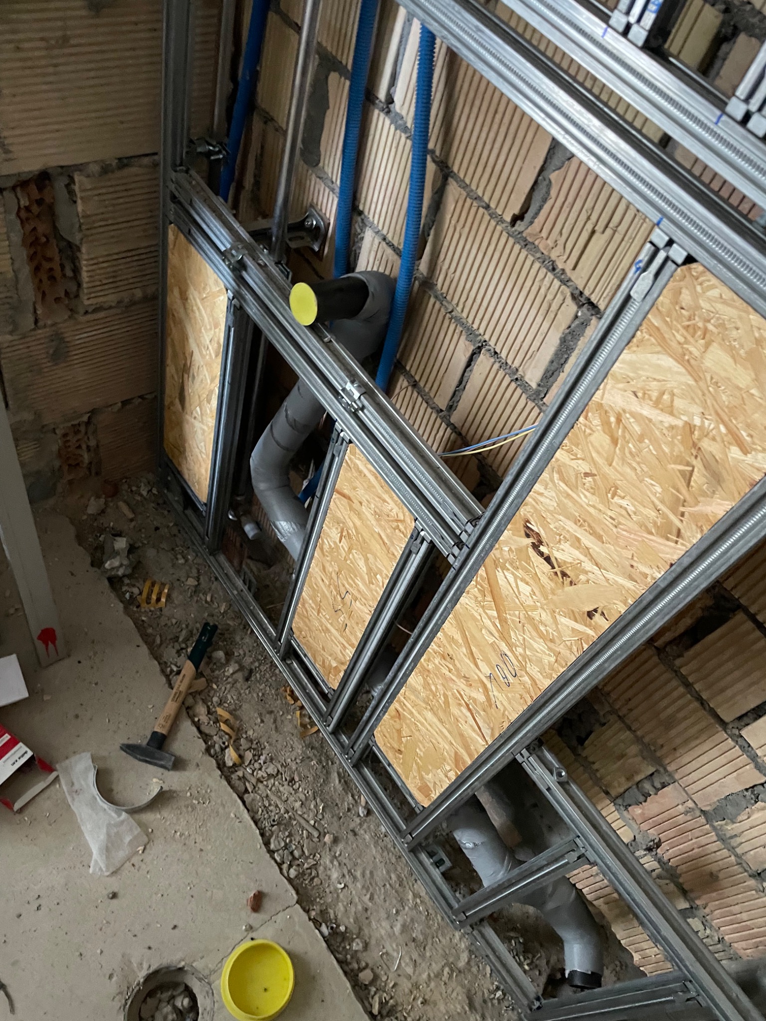 Wewnętrzna instalacja wodno-kanalizacyjna w trakcie montażu: widoczne rury PCV, metalowy stelaż, płyty OSB, fragment ceglanej ściany i podłoga w stanie surowym z narzędziami.