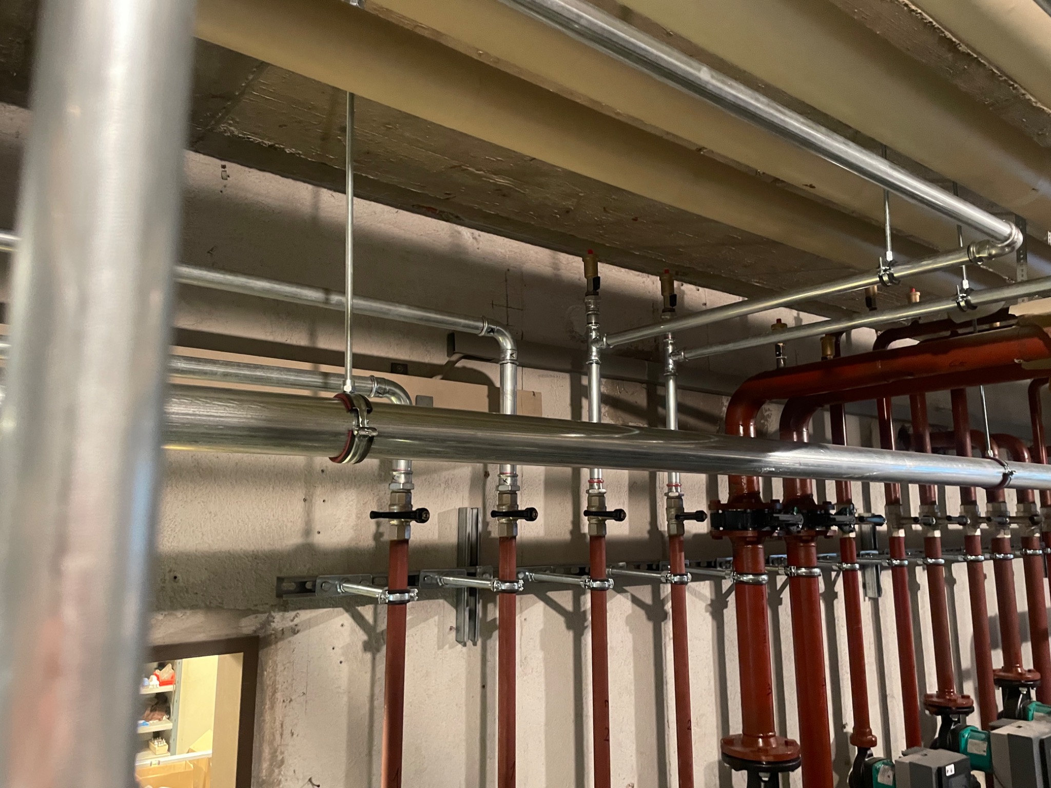 Skomplikowany system rur grzewczych w piwnicy, z widocznymi zaworami i pompami, oświetlony sztucznym światłem, z fragmentem regału widocznym w tle przez otwarte drzwi.