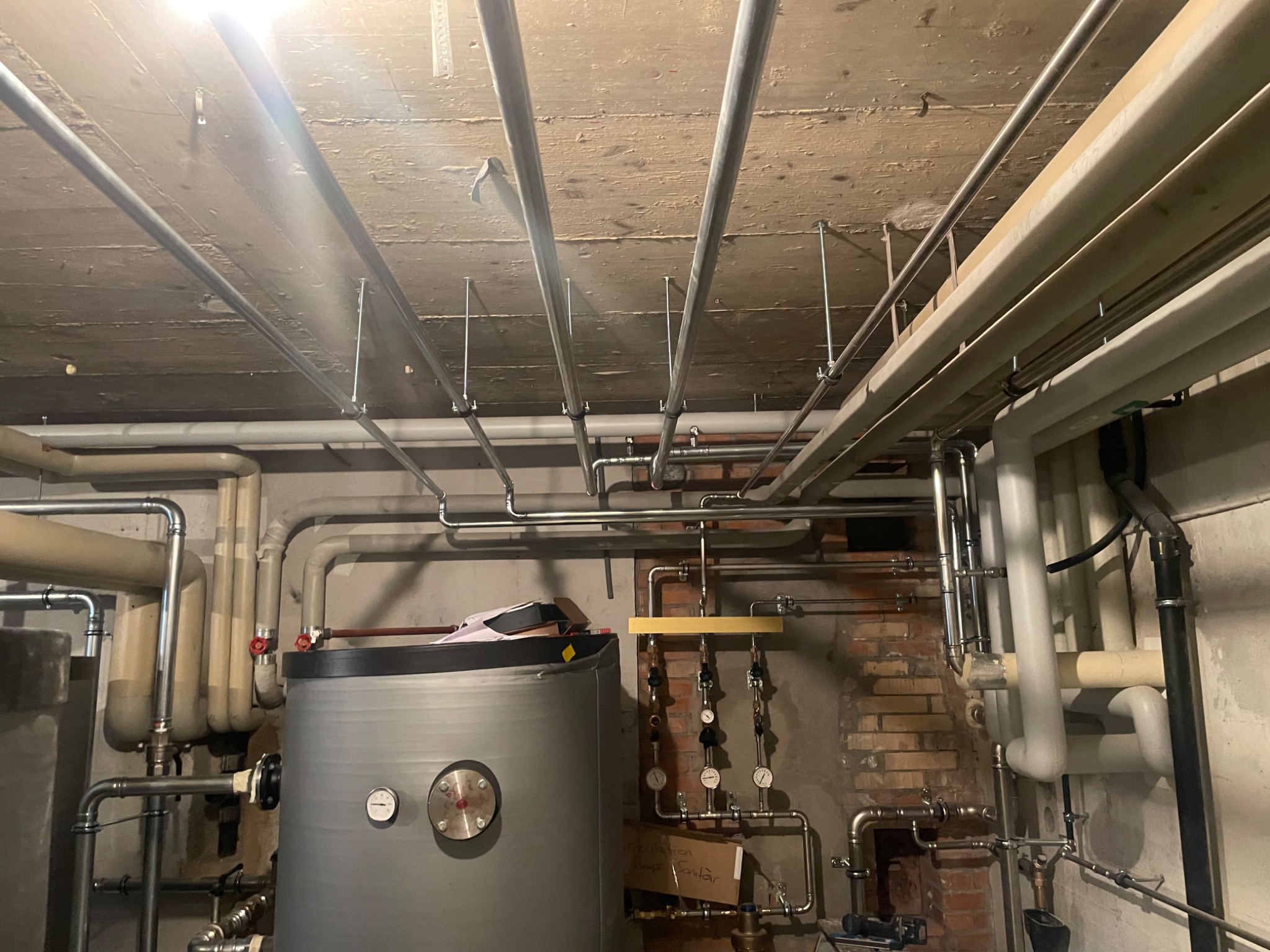 Kotłownia z widocznymi rurami instalacji grzewczej i sanitarnej, zbiornikiem akumulacyjnym oraz fragmentem ściany z cegły, oświetlona jasnym światłem.