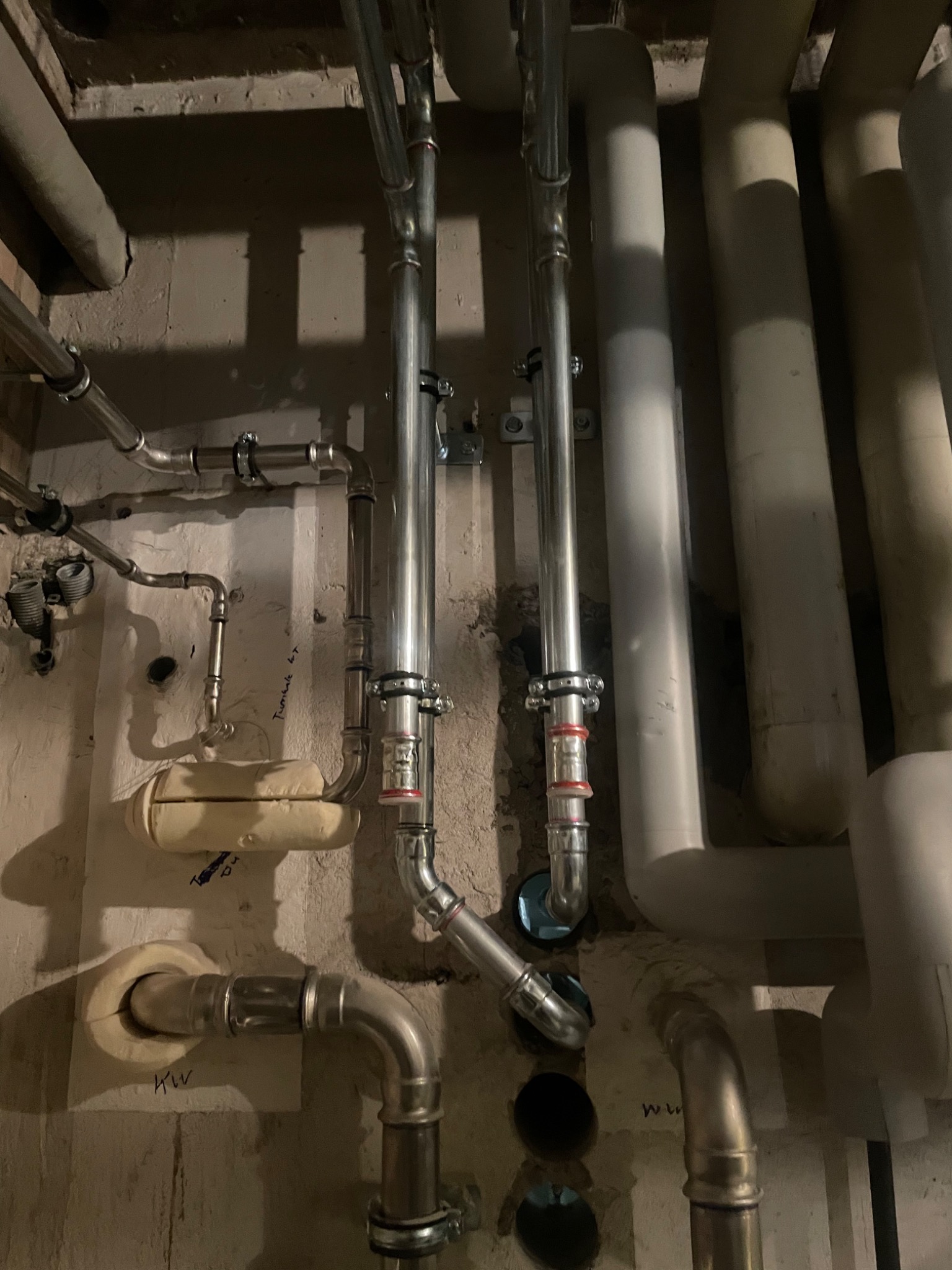 Szczegółowe ujęcie instalacji hydraulicznej z rurami stalowymi o różnych średnicach, widoczne złączki, izolacje, a także ślady po wierceniach w ścianie, z oznaczeniami 'KW' i 'WW' na ścianie.