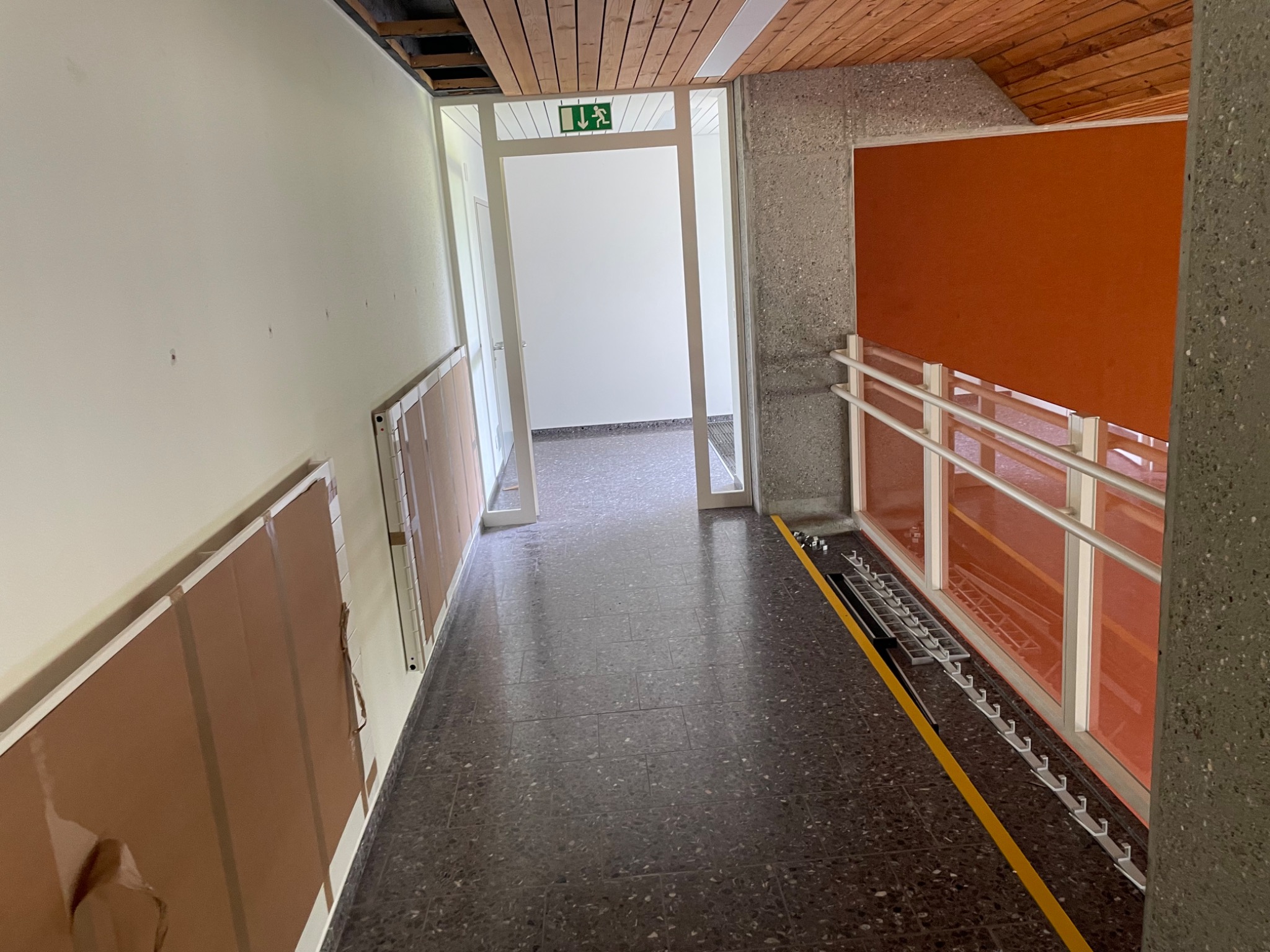 Korytarz w trakcie remontu, zabezpieczone grzejniki panelowe przy ścianie, widoczne elementy instalacji pod sufitem, drabina oparta o ścianę z pomarańczowym panelem.