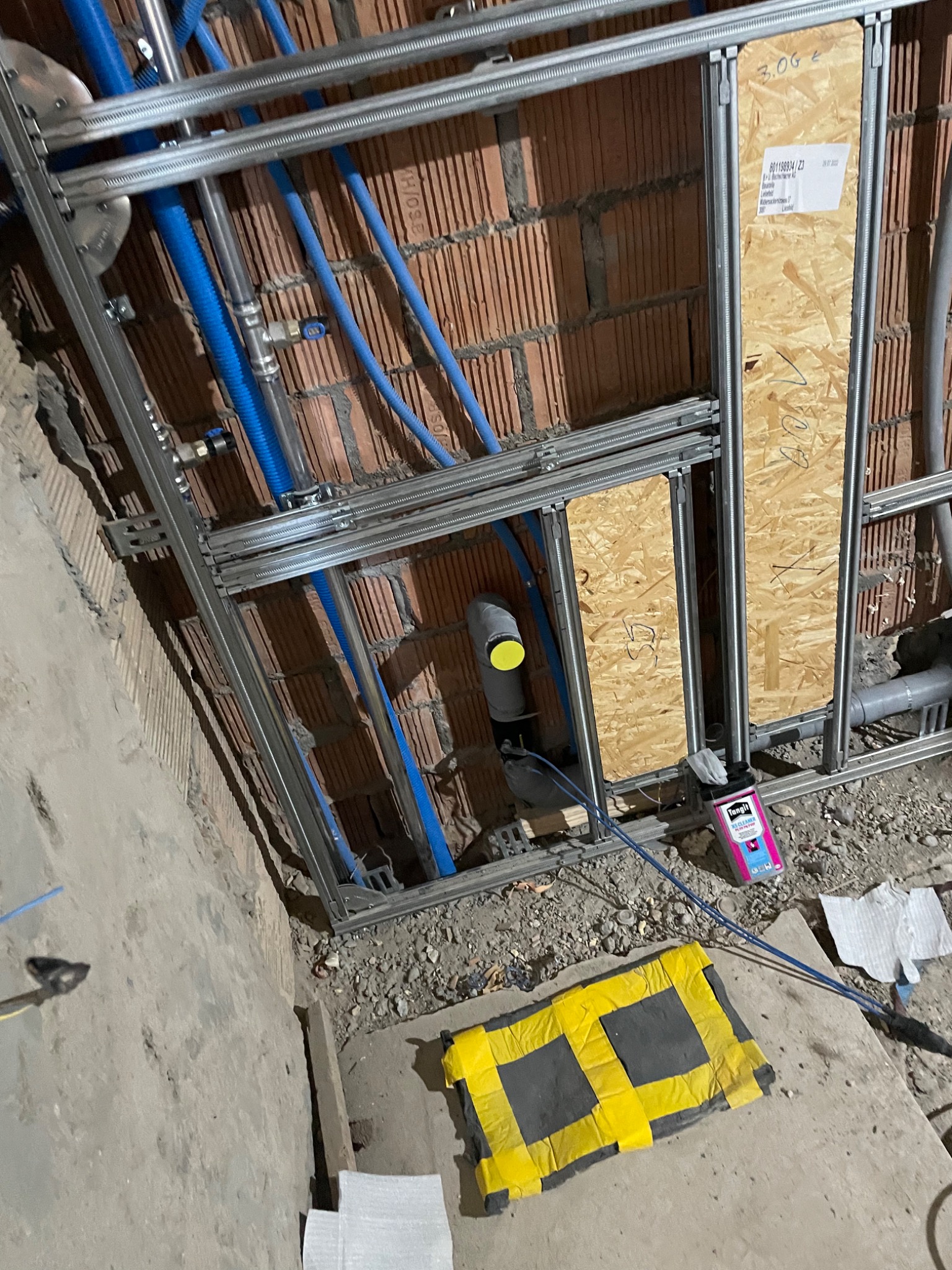 Wnętrze w trakcie montażu instalacji wodno-kanalizacyjnej z widocznymi niebieskimi rurami, szarą konstrukcją metalową mocującą oraz fragmentem ściany z cegły i płyty OSB z notatkami. Na podłodze...