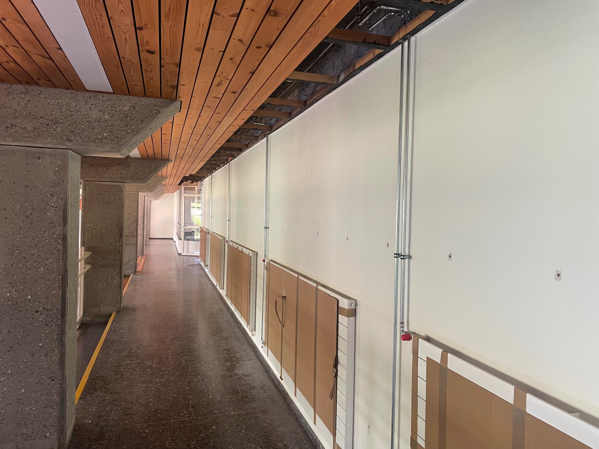 Korytarz w budynku z widocznymi nowymi instalacjami grzewczymi, grzejniki zabezpieczone kartonem, rury prowadzone wzdłuż ściany.