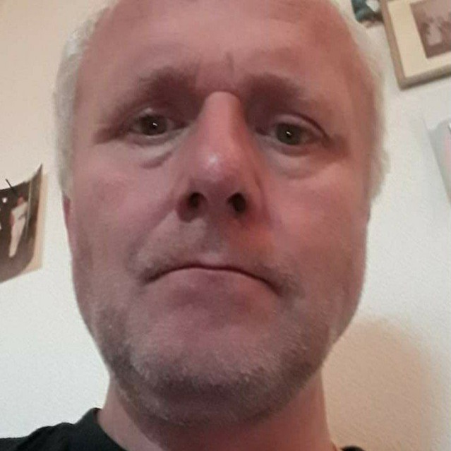 Portret mężczyzny z siwiejącymi włosami i lekkim zarostem, zarysowanymi zmarszczkami na twarzy, patrzącego wprost w obiektyw, z elementami dekoracyjnymi na ścianie w tle.