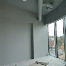 Wnętrze biura w trakcie prac wykończeniowych: ściany pomalowane na szaro, widoczne instalacje pod sufitem oraz materiały budowlane na podłodze przy oknie.