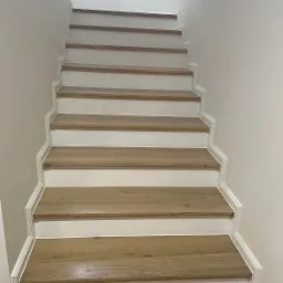 Schody wewnętrzne z drewnianymi stopniami i białymi podstopnicami, wykończone listwami przypodłogowymi o profilowanym kształcie.