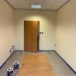 Biuro w trakcie remontu: częściowo ułożone panele podłogowe w odcieniach brązu, ściany przygotowane do malowania z widocznymi puszkami farb Dekoral i wałkiem malarskim, zabezpieczone folią i taśmą...