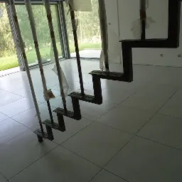 Wnętrze w trakcie wykańczania: białe płytki podłogowe, minimalistyczne, czarne schody bez balustrady zabezpieczone folią, duże okno z widokiem na zieleń.