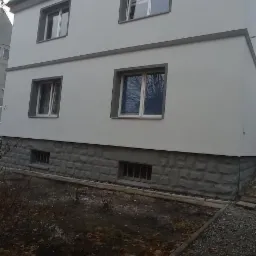 Dwupiętrowy budynek po remoncie elewacji, z szarą fasadą i ciemniejszymi obramowaniami okien, widoczne jesienne liście na ziemi.