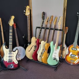 RB Music Radosław Bednarek - Kolekcja gitar elektrycznych i basowych różnych marek i kolorów, ustawionych na stojakach i podłodze w pomieszczeniu z panelami akustycznymi.