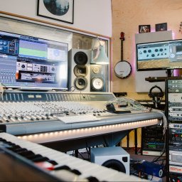 RB Music Radosław Bednarek - Profesjonalne studio nagrań: mikser dźwięku, monitory studyjne, instrumenty (banjo, keyboard) i rack ze sprzętem audio. Widoczne ekrany z oprogramowaniem DAW.