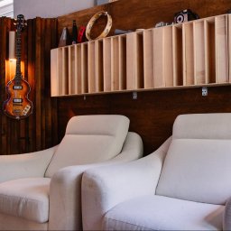 RB Music Radosław Bednarek - Wnętrze studia nagrań: dwa białe fotele, gitara basowa na ścianie, półka z instrumentami i mikrofonem. Drewniane panele akustyczne w tle.