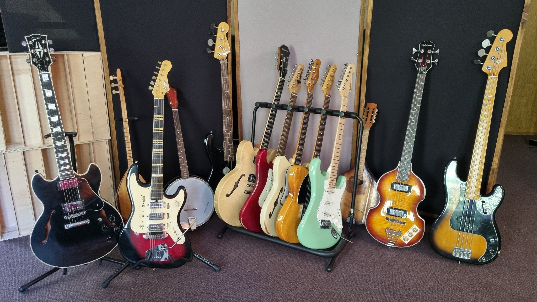 Kolekcja gitar elektrycznych i basowych różnych marek i kolorów, ustawionych na stojakach i podłodze w pomieszczeniu z panelami akustycznymi.