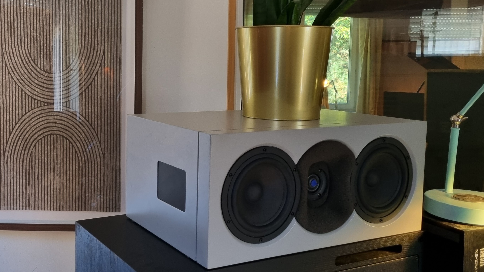 Biały głośnik trójdrożny z doniczką na górze, obok lampka i grafika na ścianie. Sprzęt audio w domowym otoczeniu, potencjalnie w studio nagrań.