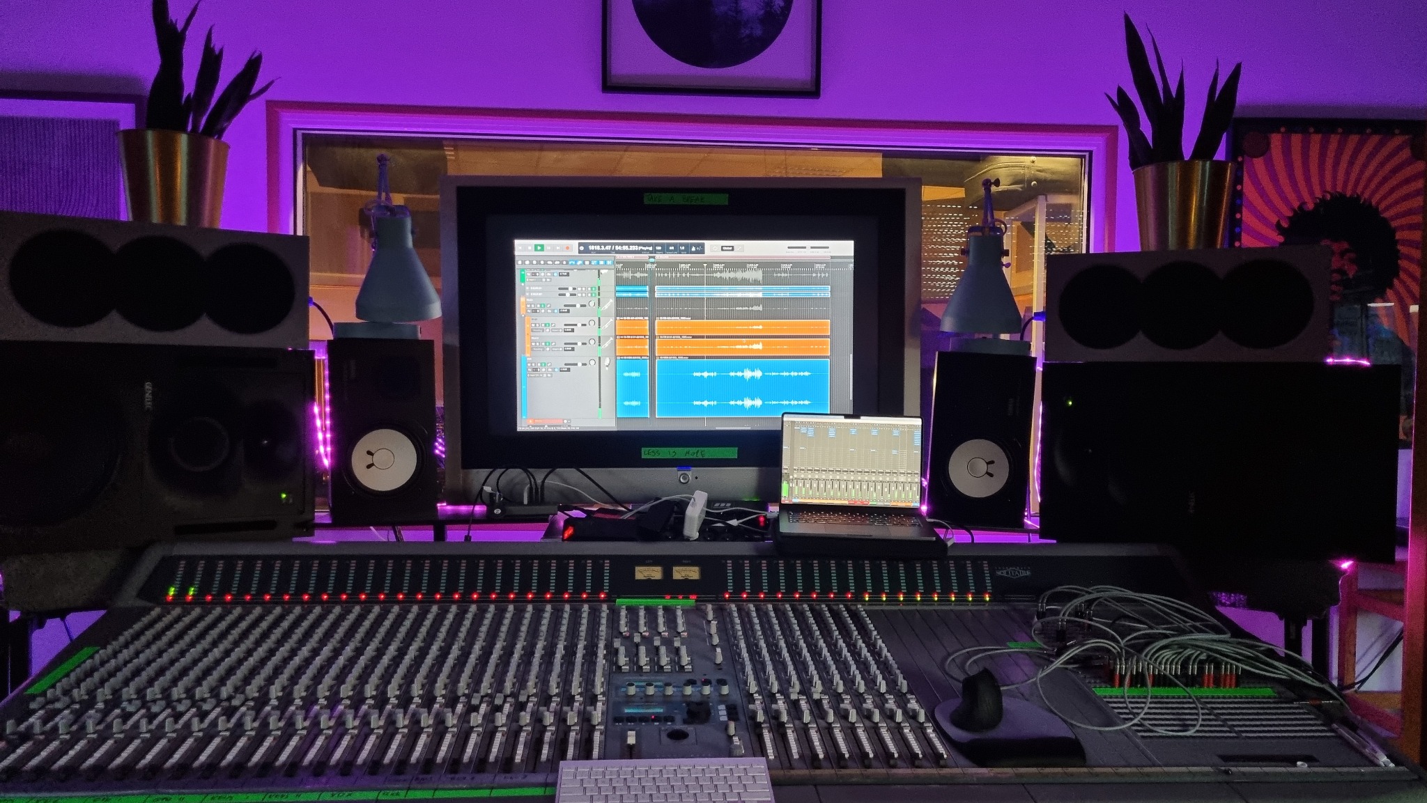 Profesjonalne studio nagrań z mikserem, monitorami studyjnymi i ekranem z oprogramowaniem DAW, oświetlone fioletowym światłem. Widoczne rośliny doniczkowe i grafika na ścianie.