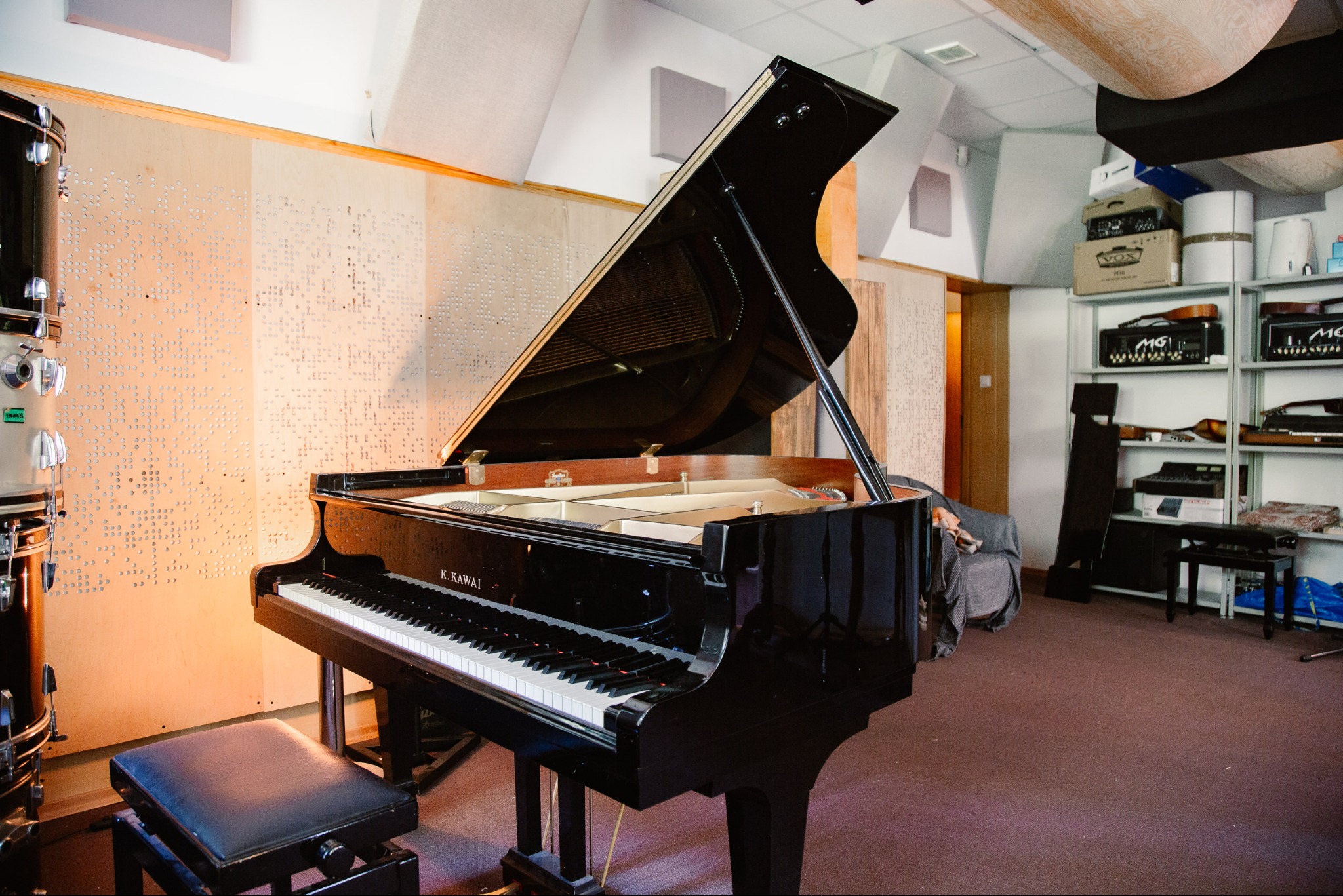 Czarny fortepian K. Kawai w profesjonalnym studio nagrań z panelami akustycznymi i perkusją. Widoczne wzmacniacze gitarowe na półkach.