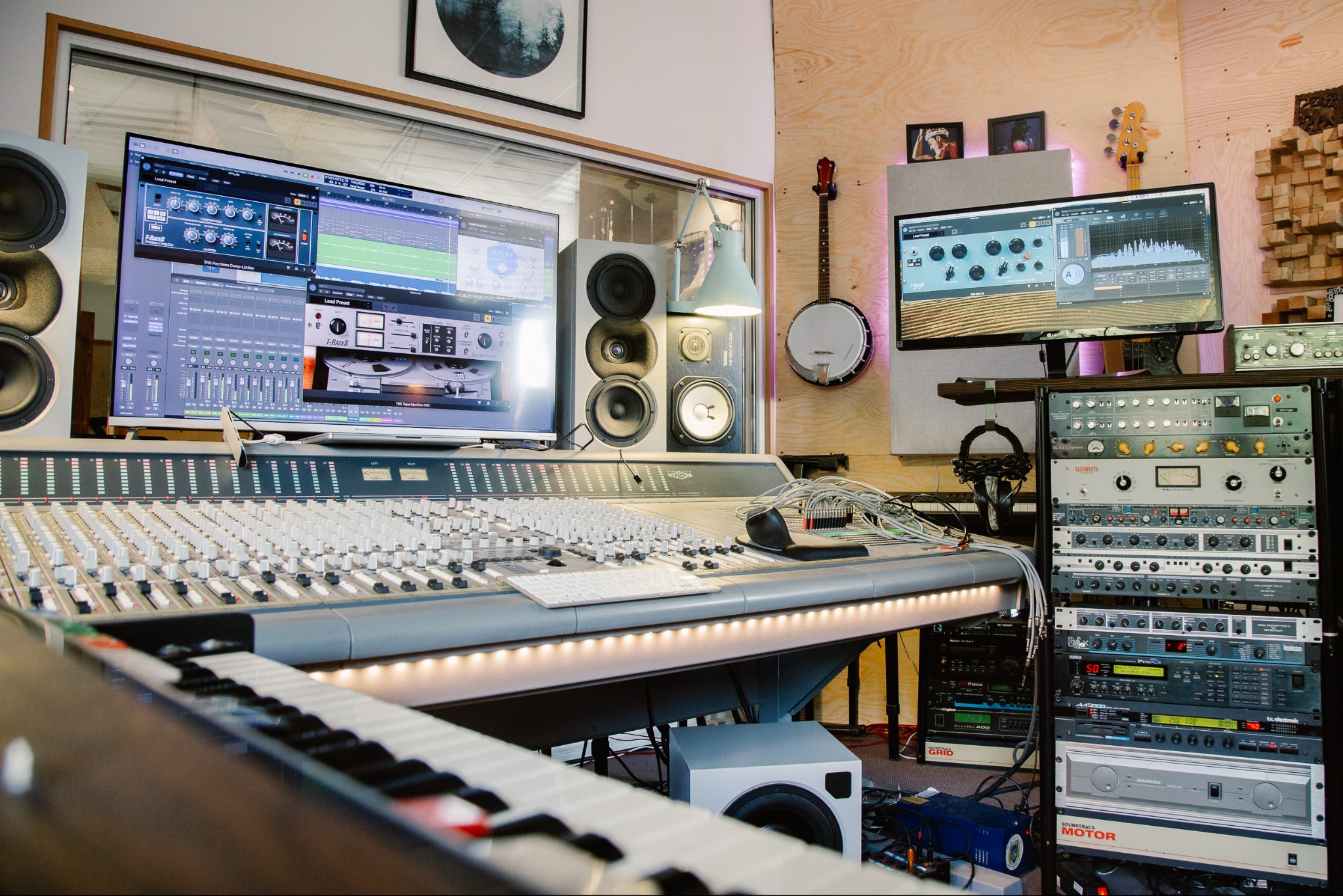 Profesjonalne studio nagrań: mikser dźwięku, monitory studyjne, instrumenty (banjo, keyboard) i rack ze sprzętem audio. Widoczne ekrany z oprogramowaniem DAW.