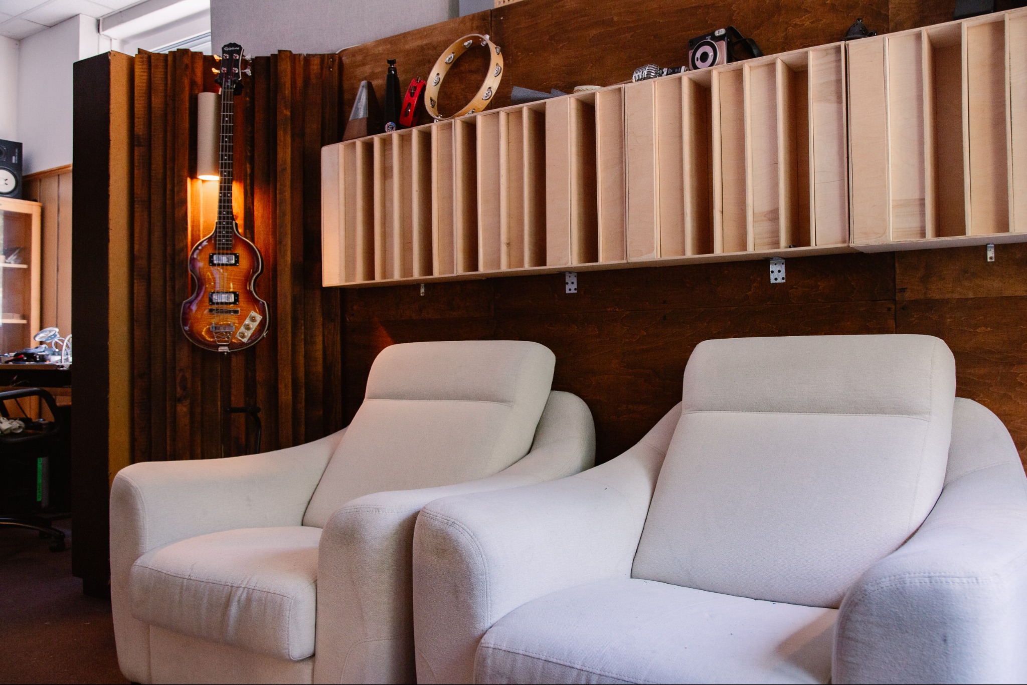 Wnętrze studia nagrań: dwa białe fotele, gitara basowa na ścianie, półka z instrumentami i mikrofonem. Drewniane panele akustyczne w tle.