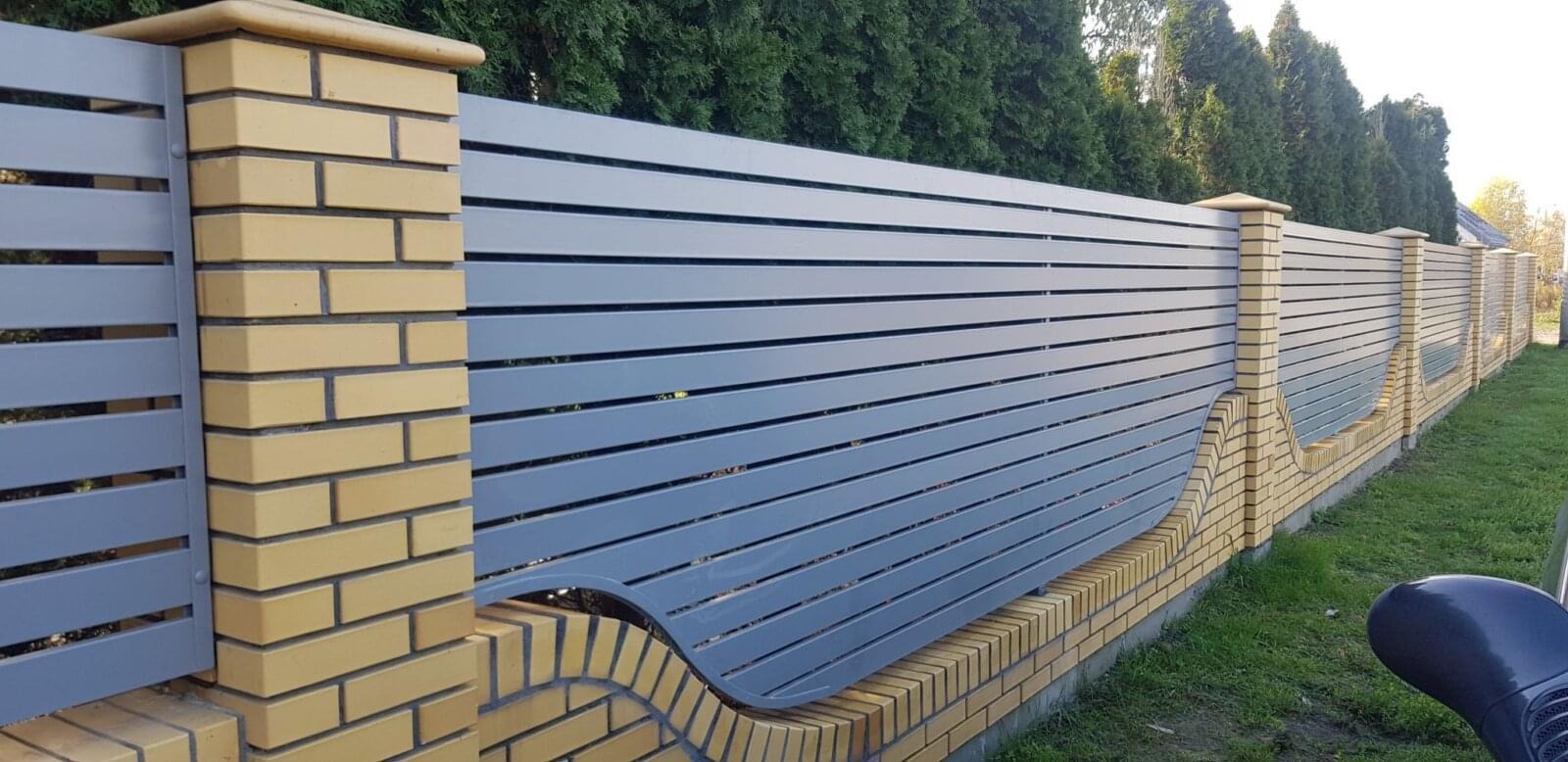 Nowoczesne ogrodzenie z poziomych, szarych desek i słupków z żółtej cegły klinkierowej z dekoracyjnym, falistym wycięciem u dołu, na tle zielonych tuj.
