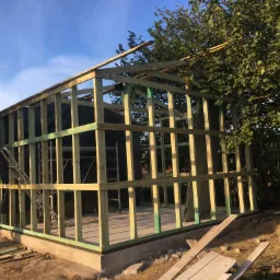 Konstrukcja szkieletowa budynku w trakcie budowy, widoczne drewniane belki pomalowane na zielono, drabina oparta o ścianę, teren budowy z piaskiem i resztkami materiałów.