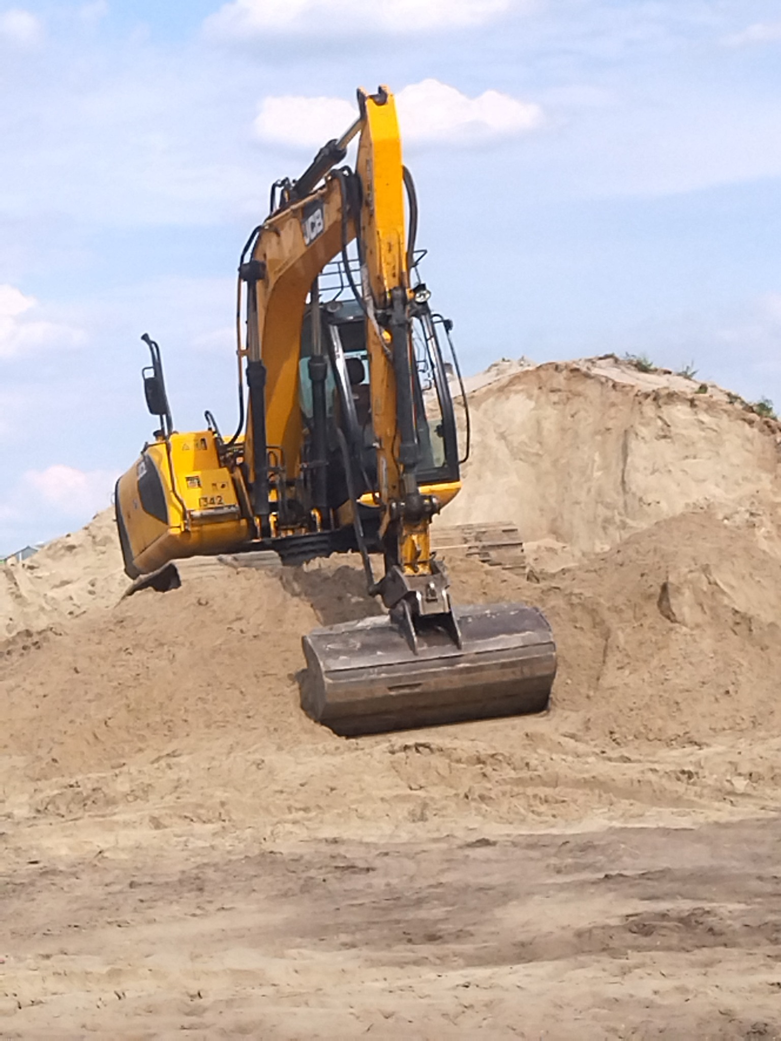 Żółta koparka JCB na szczycie piaszczystej pryzmy, widoczny numer 1342 na boku, błękitne niebo w tle.
