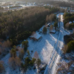 Zimowy krajobraz z lotu ptaka: ośnieżone domy, las i droga z długimi cieniami drzew.