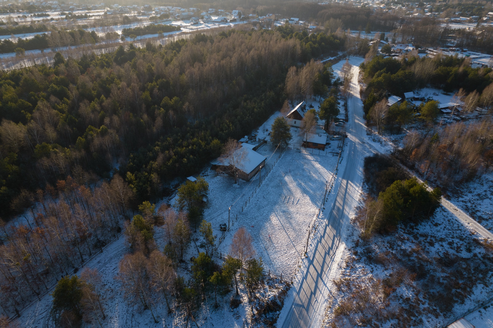 Zimowy krajobraz z lotu ptaka: ośnieżone domy, las i droga z długimi cieniami drzew.