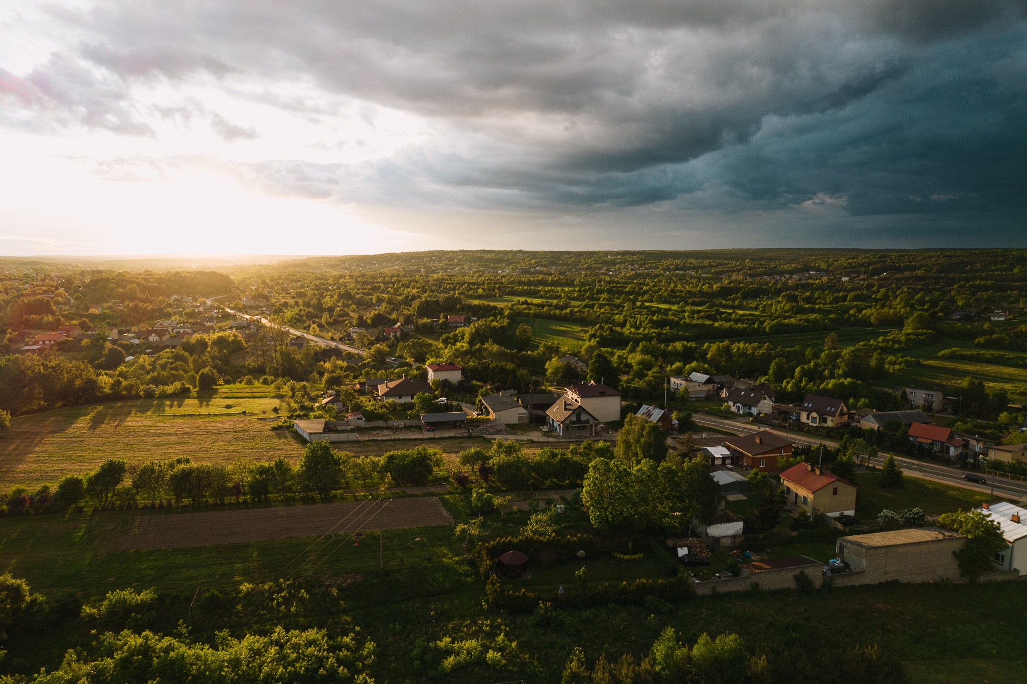 Panoramiczny widok z lotu ptaka na wiejską okolicę oświetloną zachodzącym słońcem, z ciemnymi chmurami burzowymi w tle, ukazujący domy, pola i lasy.