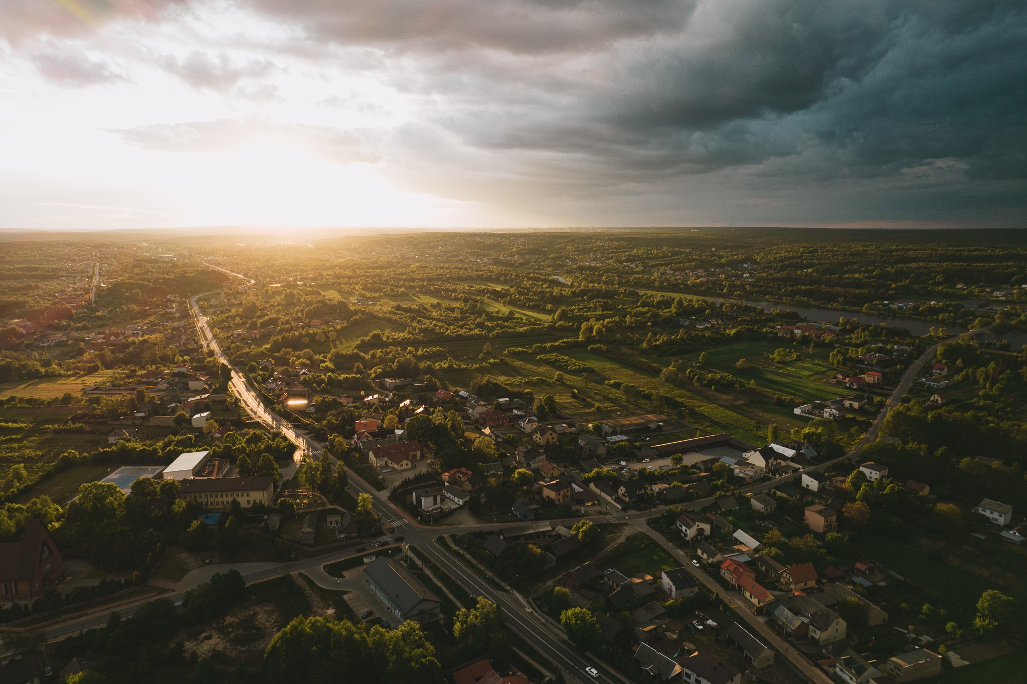 Panoramiczny widok z lotu ptaka na miasteczko i okoliczne lasy w promieniach zachodzącego słońca, z ciemnymi chmurami burzowymi w tle.