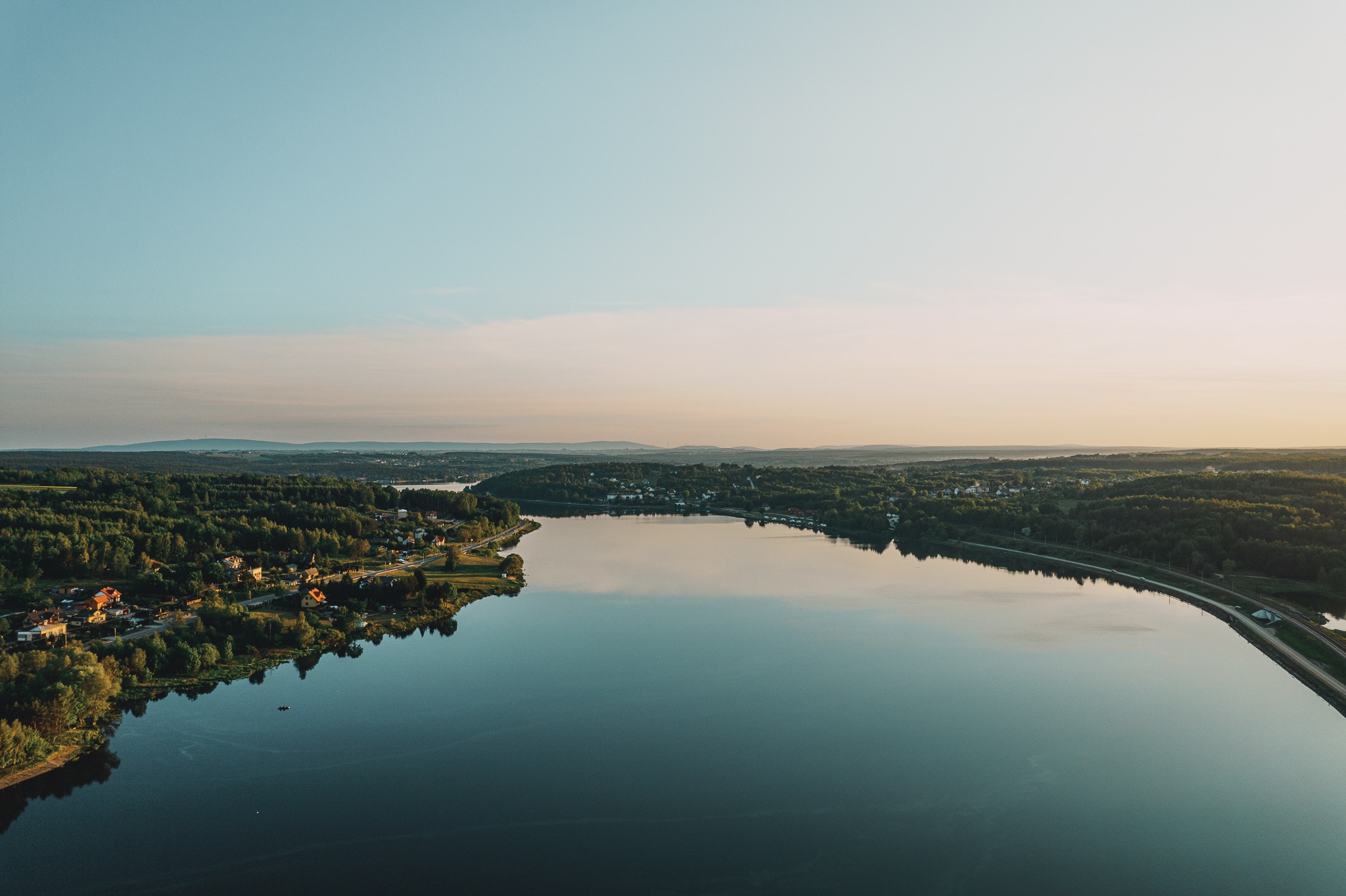 Panoramiczny widok z lotu ptaka na jezioro otoczone lasami i zabudowaniami, z delikatnym odcieniem zachodzącego słońca.