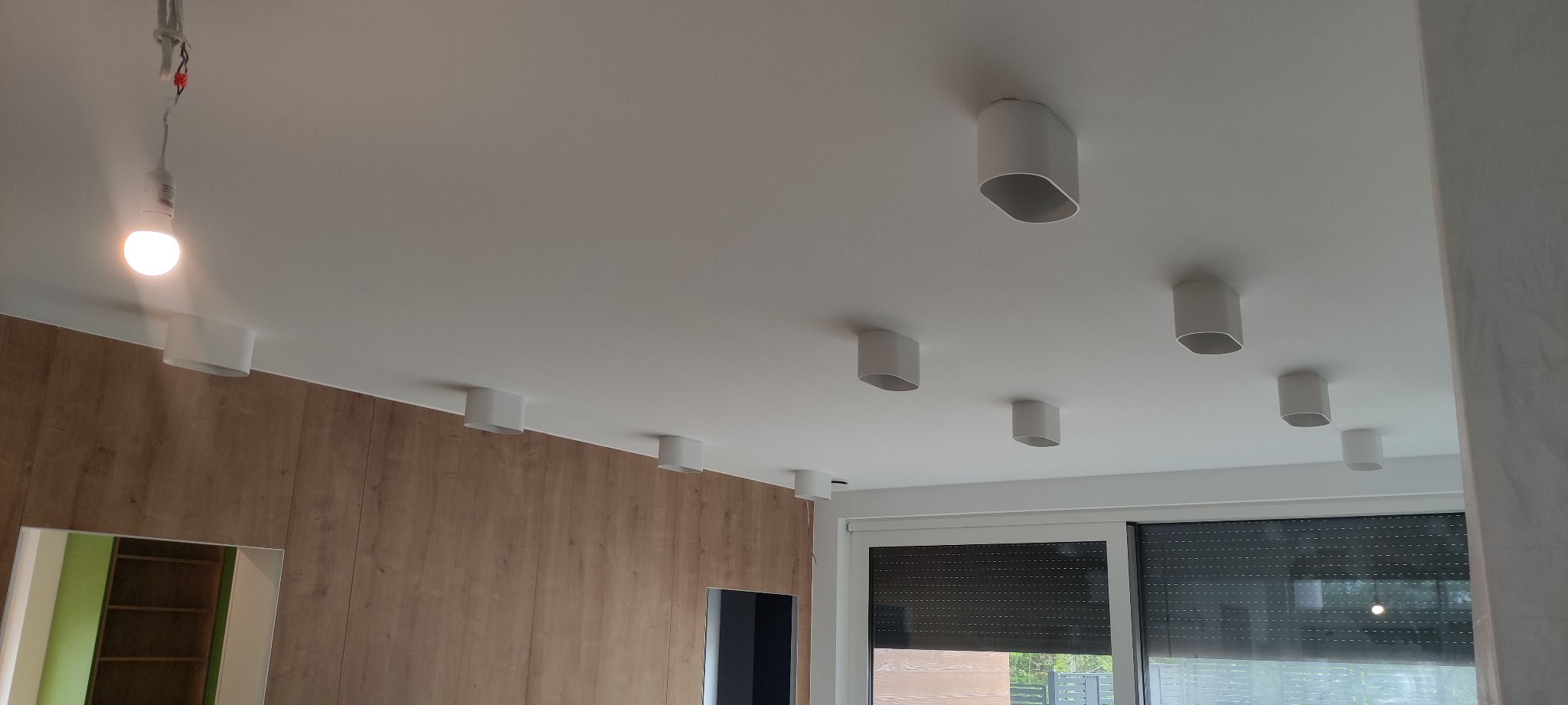 Białe, owalne lampy sufitowe zamontowane w rzędzie na białym suficie w nowoczesnym wnętrzu. Widoczna drewniana ściana i okno z żaluzjami.