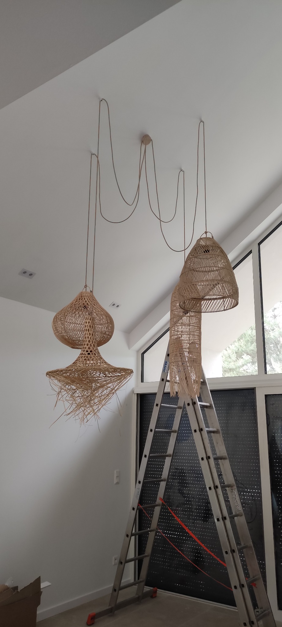 Montaż trzech plecionych lamp wiszących z długimi kablami na tle białego sufitu i drabiny. Widoczne okno z roletami.