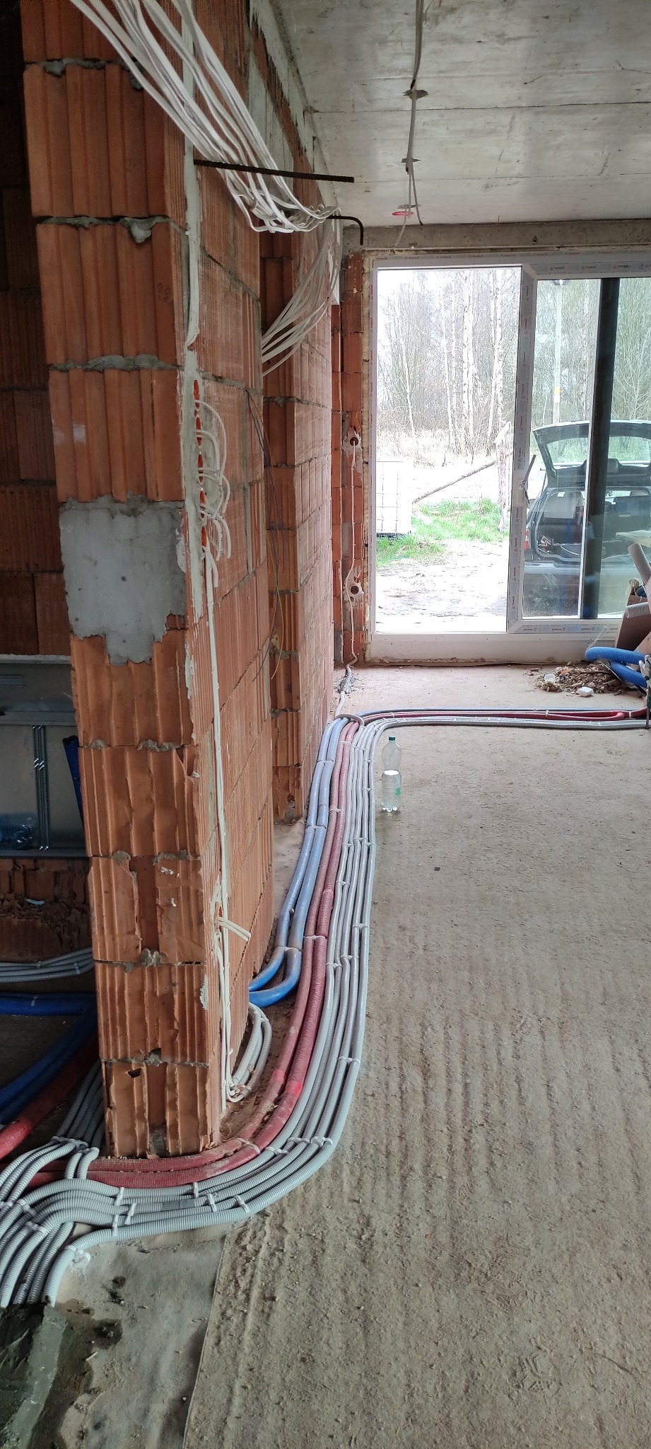 Instalacja elektryczna w surowym budynku: liczne kable prowadzone po ścianach z cegły i pod sufitem, widoczne otwory na puszki elektryczne, butelka wody na podłodze.