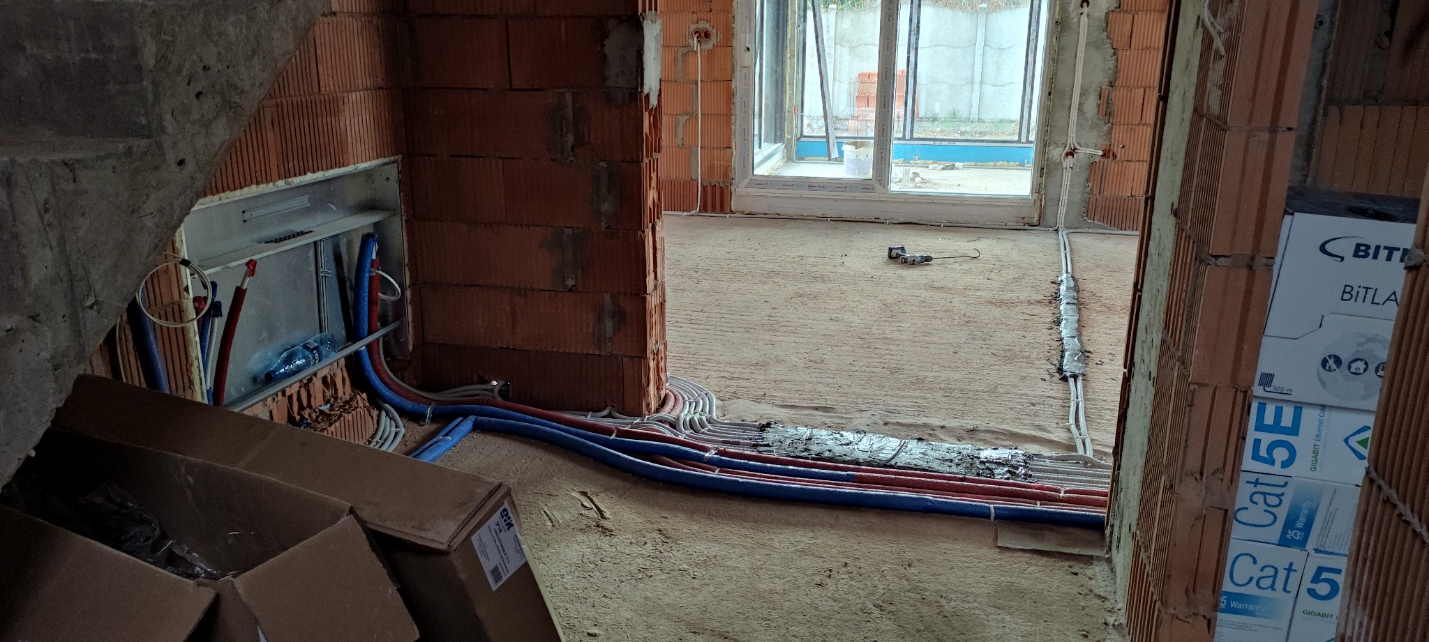 Instalacja elektryczna i hydrauliczna w surowym wnętrzu domu, widoczne przewody w ścianach, rury w podłodze oraz skrzynka rozdzielcza w przygotowanej wnęce, w tle okno z widokiem na zewnątrz.