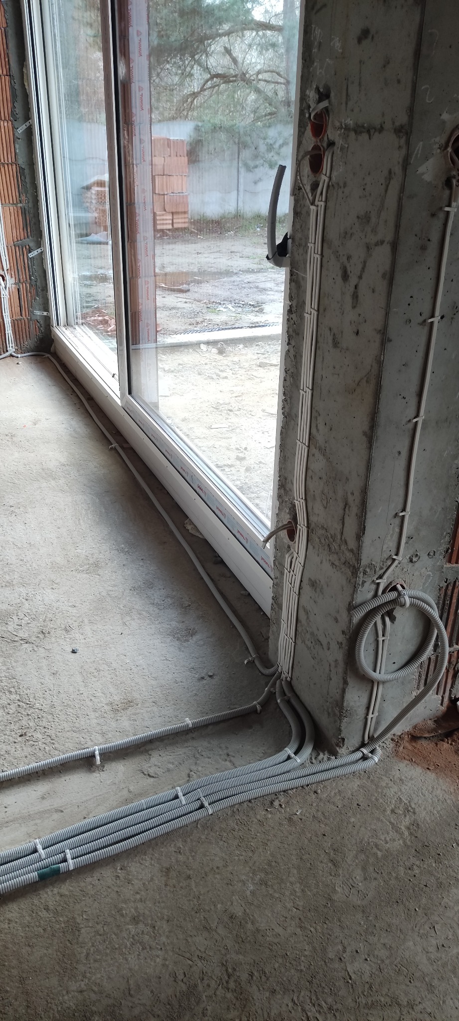 Wewnętrzne kable elektryczne w peszlach prowadzone wzdłuż betonowego filaru i podłogi w surowym stanie, widoczne otwory w ścianie i okno z widokiem na zewnątrz.