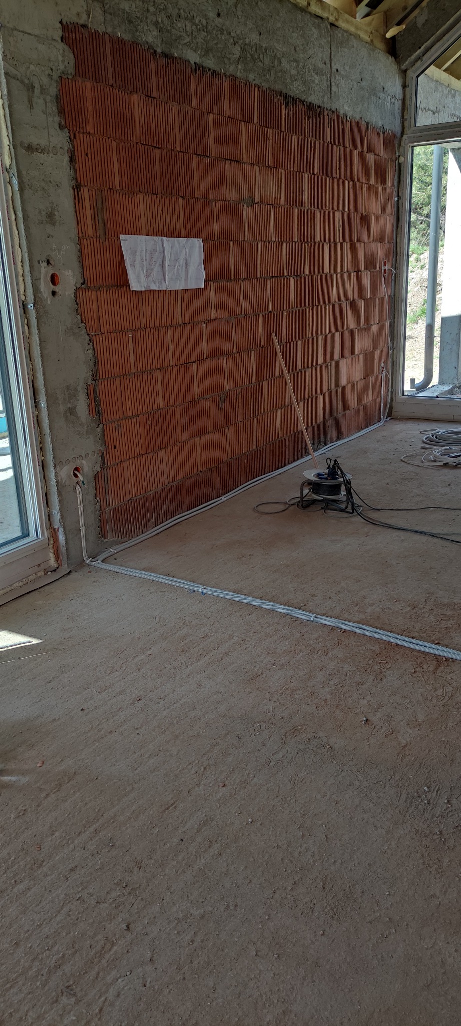 Wnętrze w trakcie prac wykończeniowych: ściana z czerwonej cegły dziurawki, częściowo otynkowana, widoczne przewody elektryczne w białych listwach przypodłogowych, rozdzielnica elektryczna...