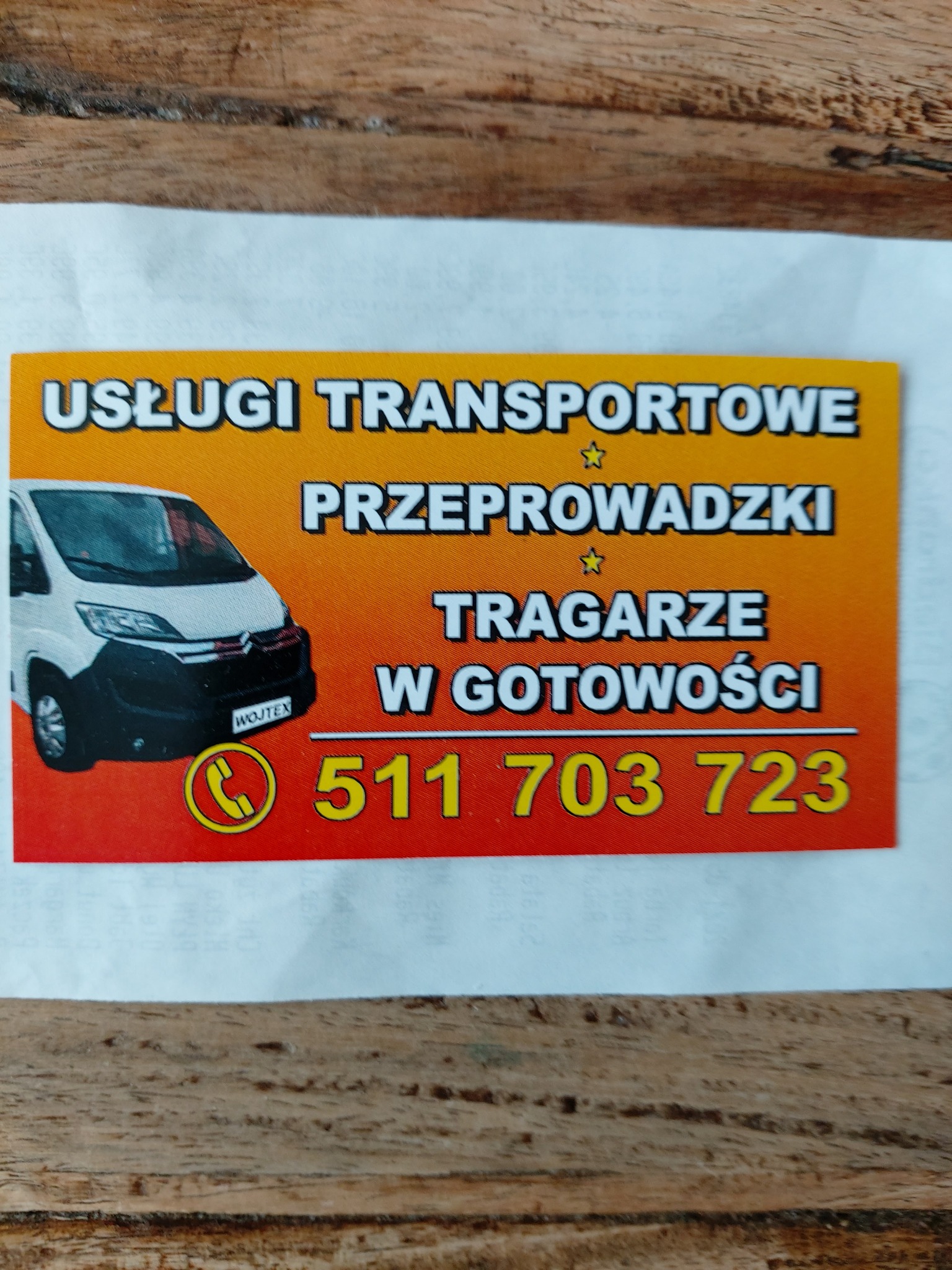Wizytówka firmy transportowej z białym busem, numerem telefonu i informacją o usługach przeprowadzkowych oraz tragarzach w gotowości na pomarańczowo-czerwonym tle.