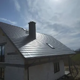 Nowy, ciemnoszary dach z dachówki ceramicznej na domu w trakcie budowy, widoczny komin, okno dachowe i rynna, w tle błękitne niebo z chmurami i fragment krajobrazu.