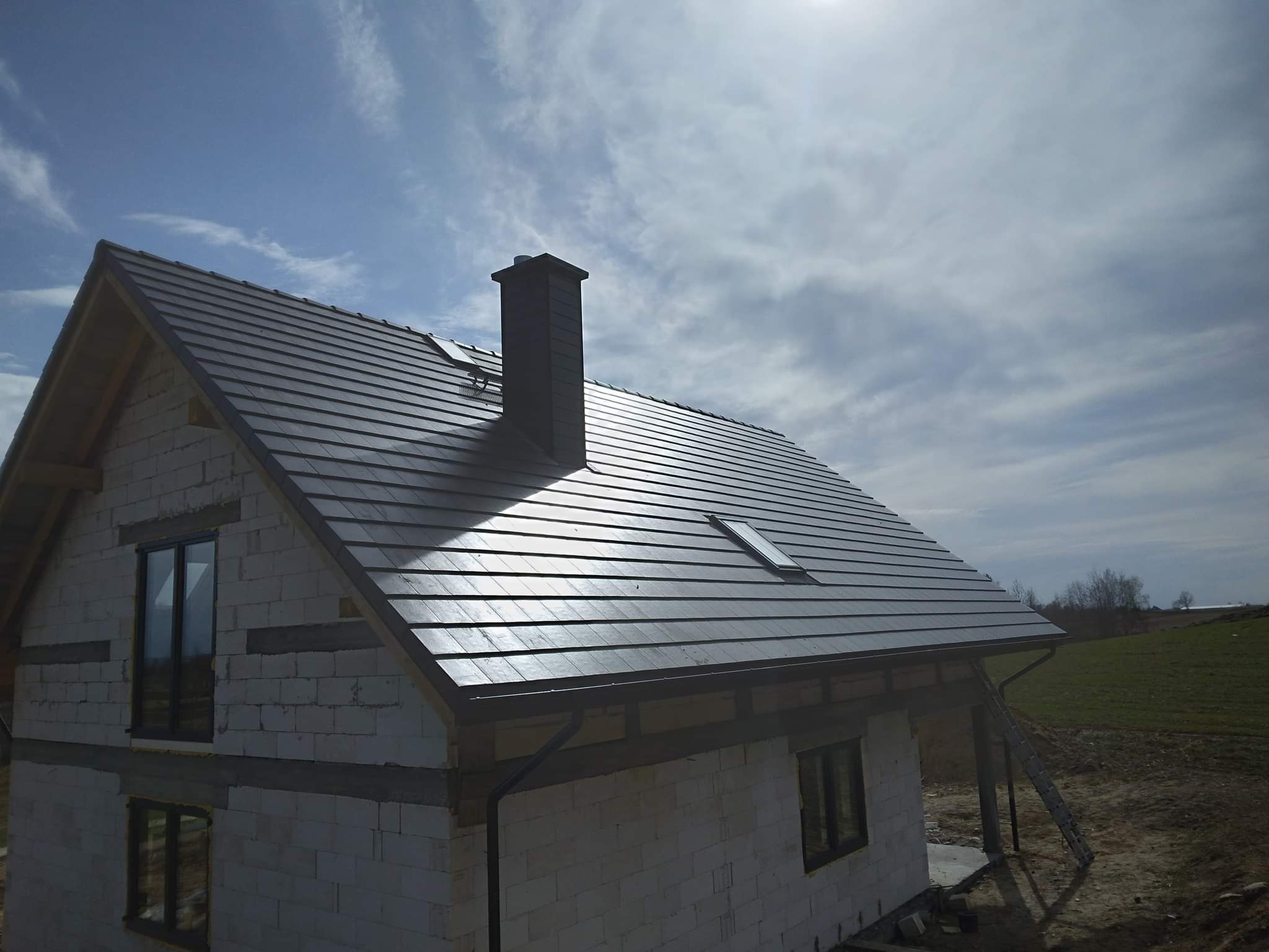 Nowy, ciemnoszary dach z dachówki ceramicznej na domu w trakcie budowy, widoczny komin, okno dachowe i rynna, w tle błękitne niebo z chmurami i fragment krajobrazu.