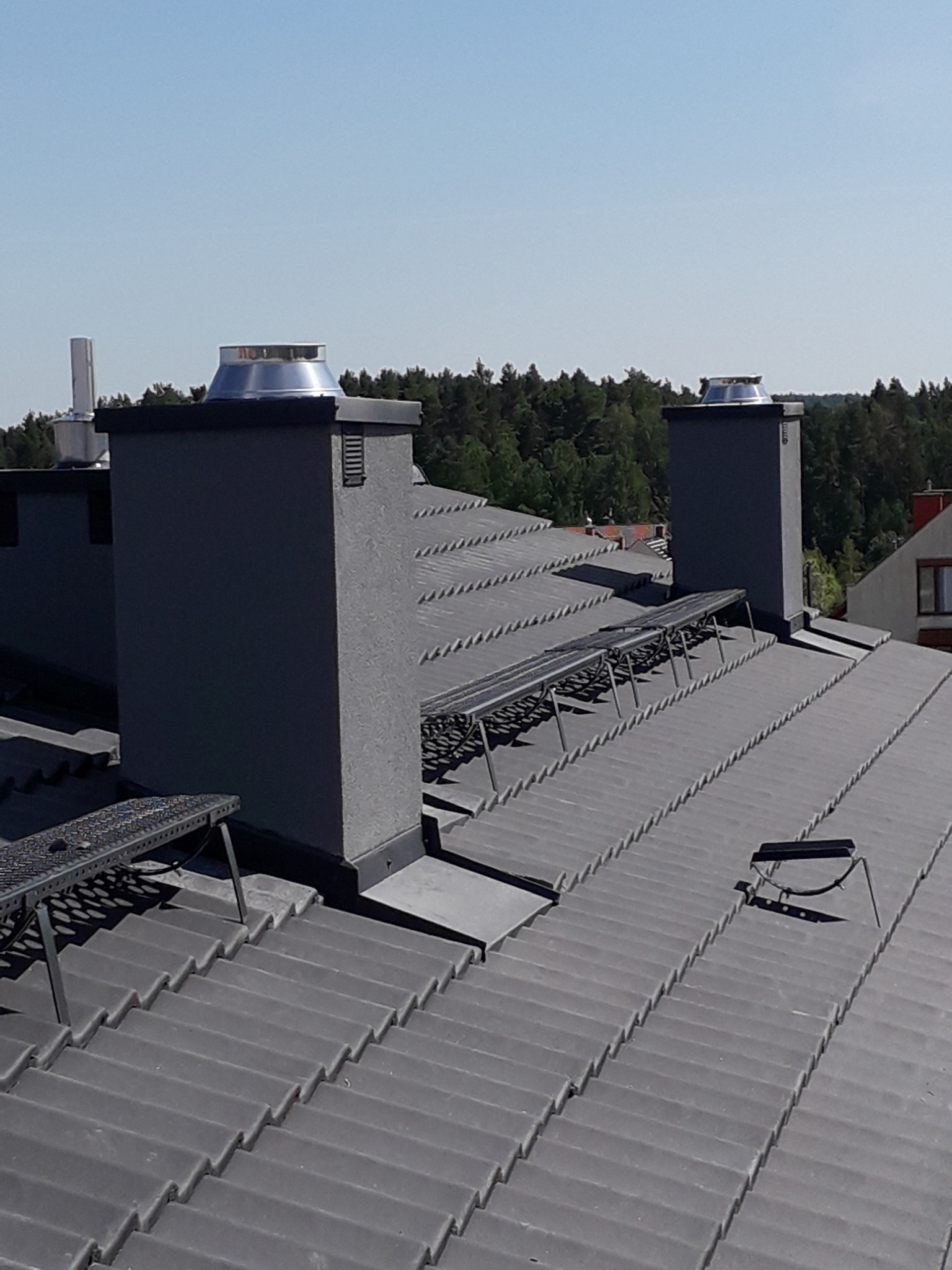 Szary dach z dachówki ceramicznej z kominami i metalowymi zabezpieczeniami przeciwśniegowymi, widok z góry w słoneczny dzień.