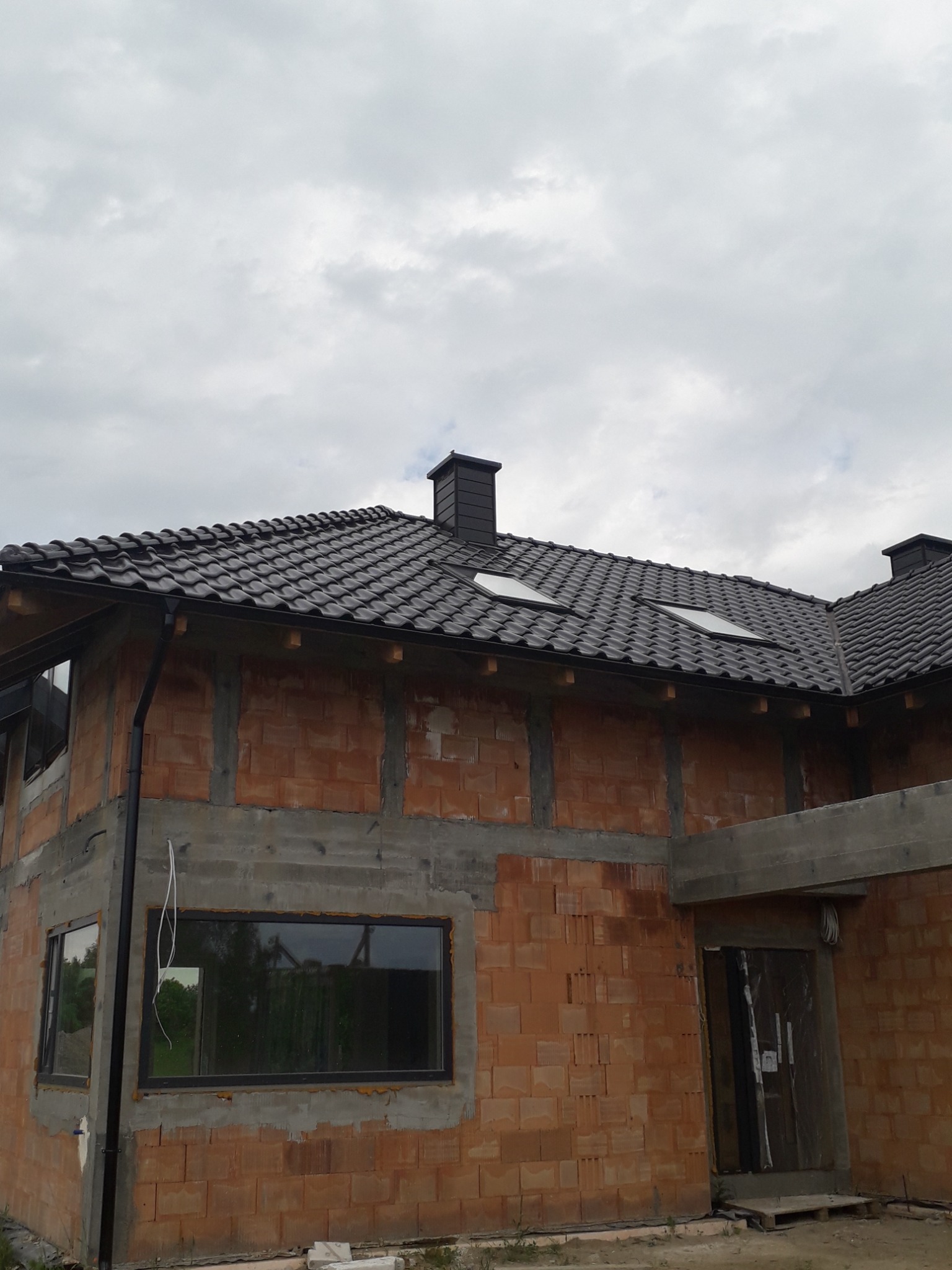 Budynek w trakcie budowy z zamontowaną, ciemną dachówką ceramiczną i oknami dachowymi; widoczne rynny, komin oraz ściany z czerwonej cegły.